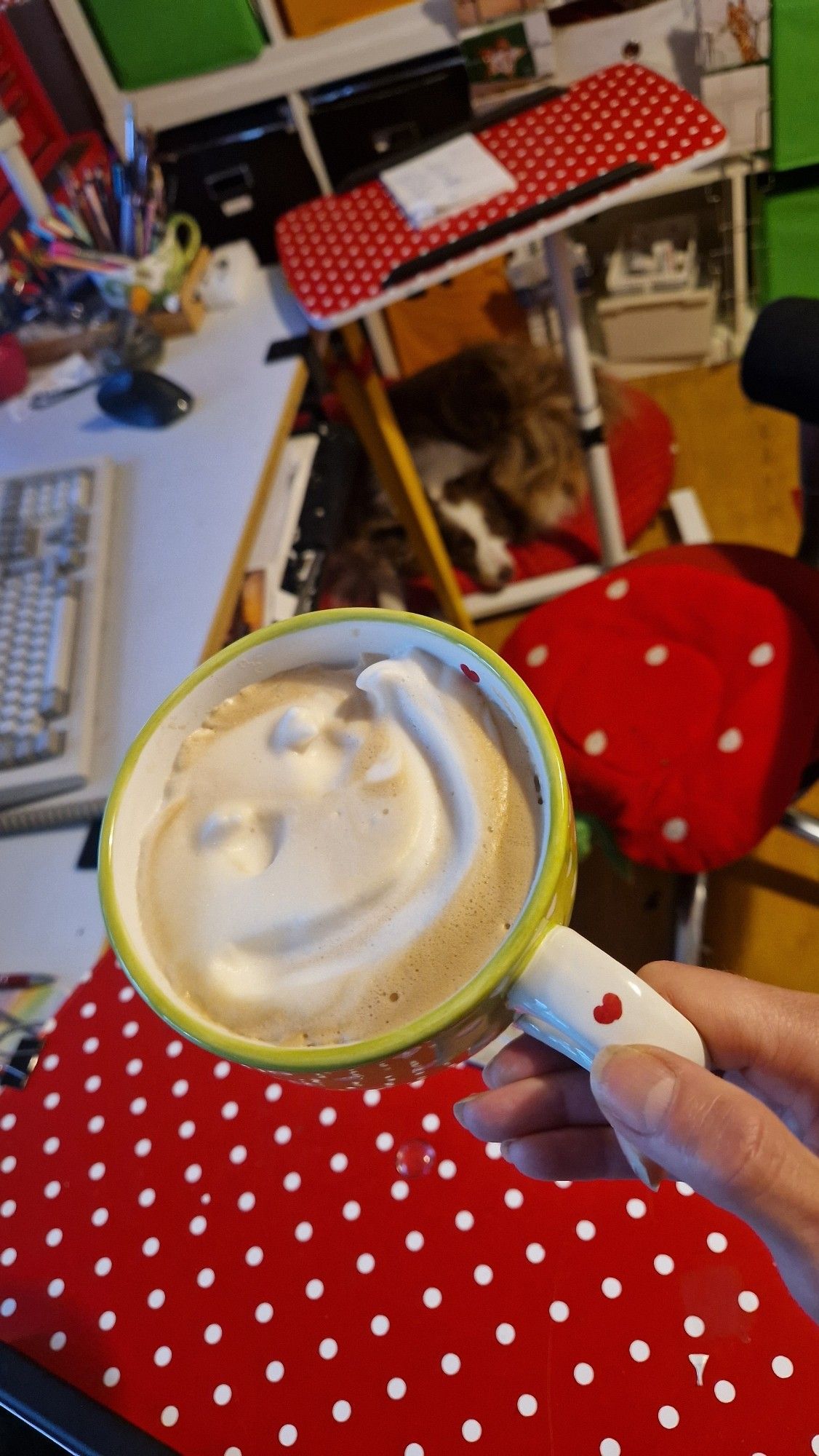 Kaffee mit smilie im Milchschaum Tasse ist mit Herzen bemalt dahinter Arbeitsplatz mit Tastatur Maus und Malsachen Am Boden auf einem Kissen liegt ein Hund