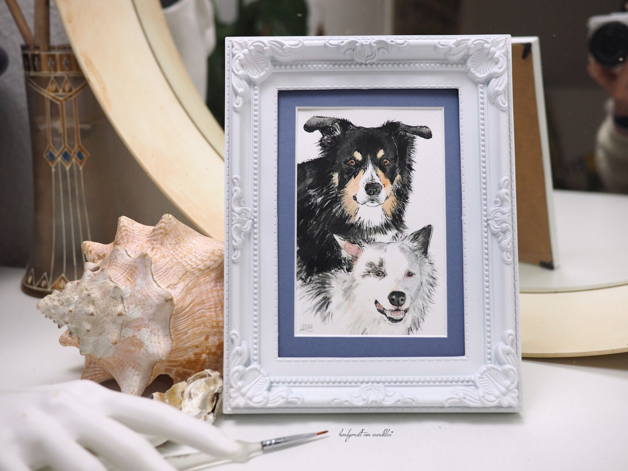 in weißen Schnörkelrahmen gefasstes Aquarell: Duo zweier Hunde, ein würdig blickender älterer tricolor Border Collie und ein durchtrieben  guckender weißer Junghund, ein (teilweise etwas graumelierter) Bordercollie-Pudel-Mix.