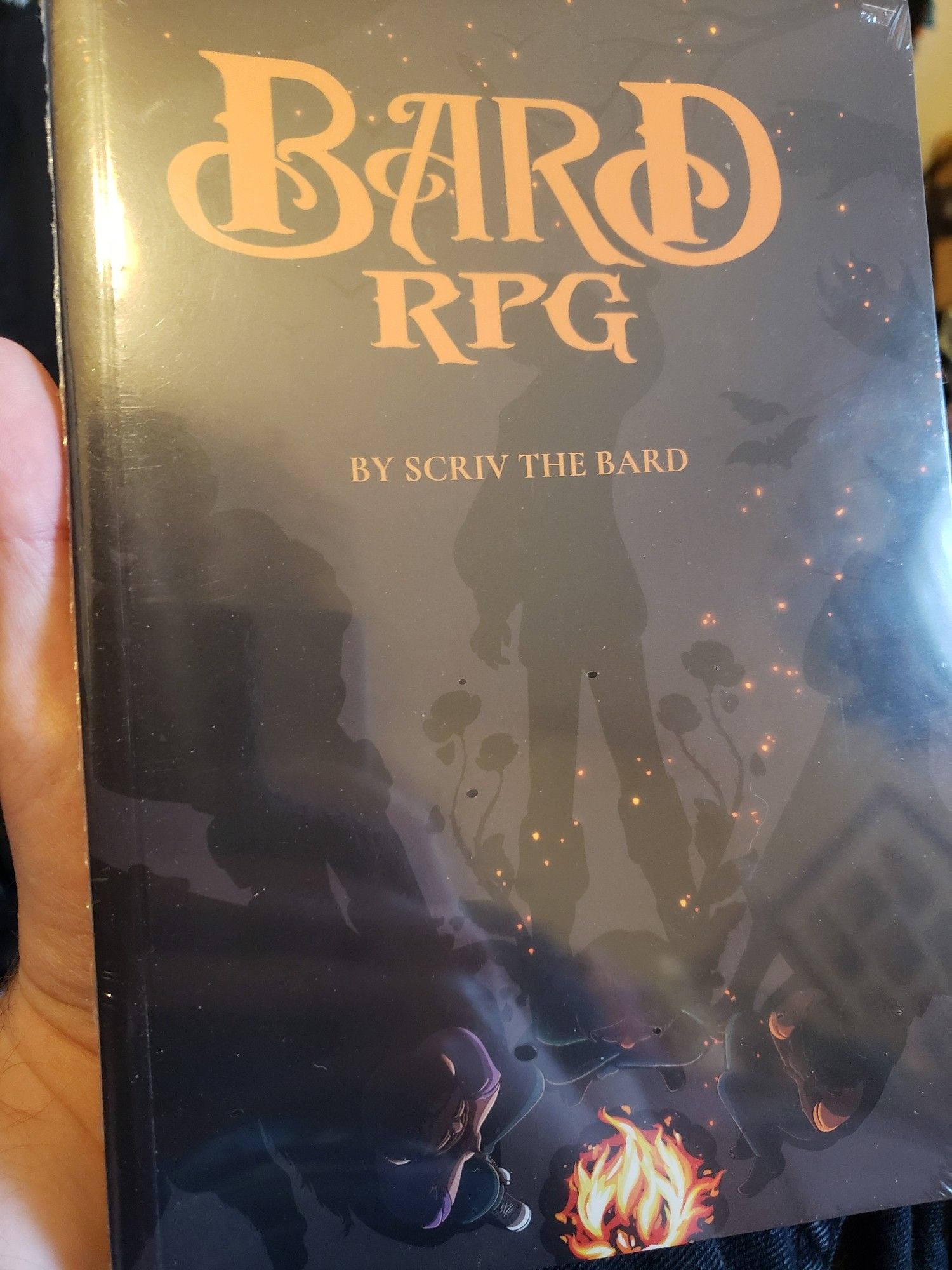 A photo of the Bardrpg book, still in wrapping. The cover shows a top down artwork of three young epople sat at a fire, their shadows extending out as fantastical characters.