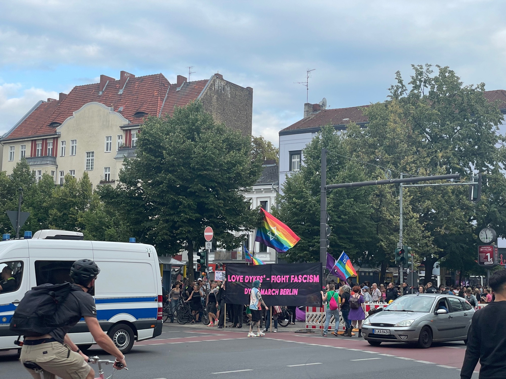 Blick über eine Kreuzung, dahinter stehen menschen und halten ein Transparent in die Höhe, darauf steht: Love Dykes* - Fight Fascism. Im Hintergrund ein Regenbogenfahne im Wind darauf ein Davidstern. Rechtsfähig ein Auto auf die Kreuzung, links ein Radfahrer und ein Polizeiauto.