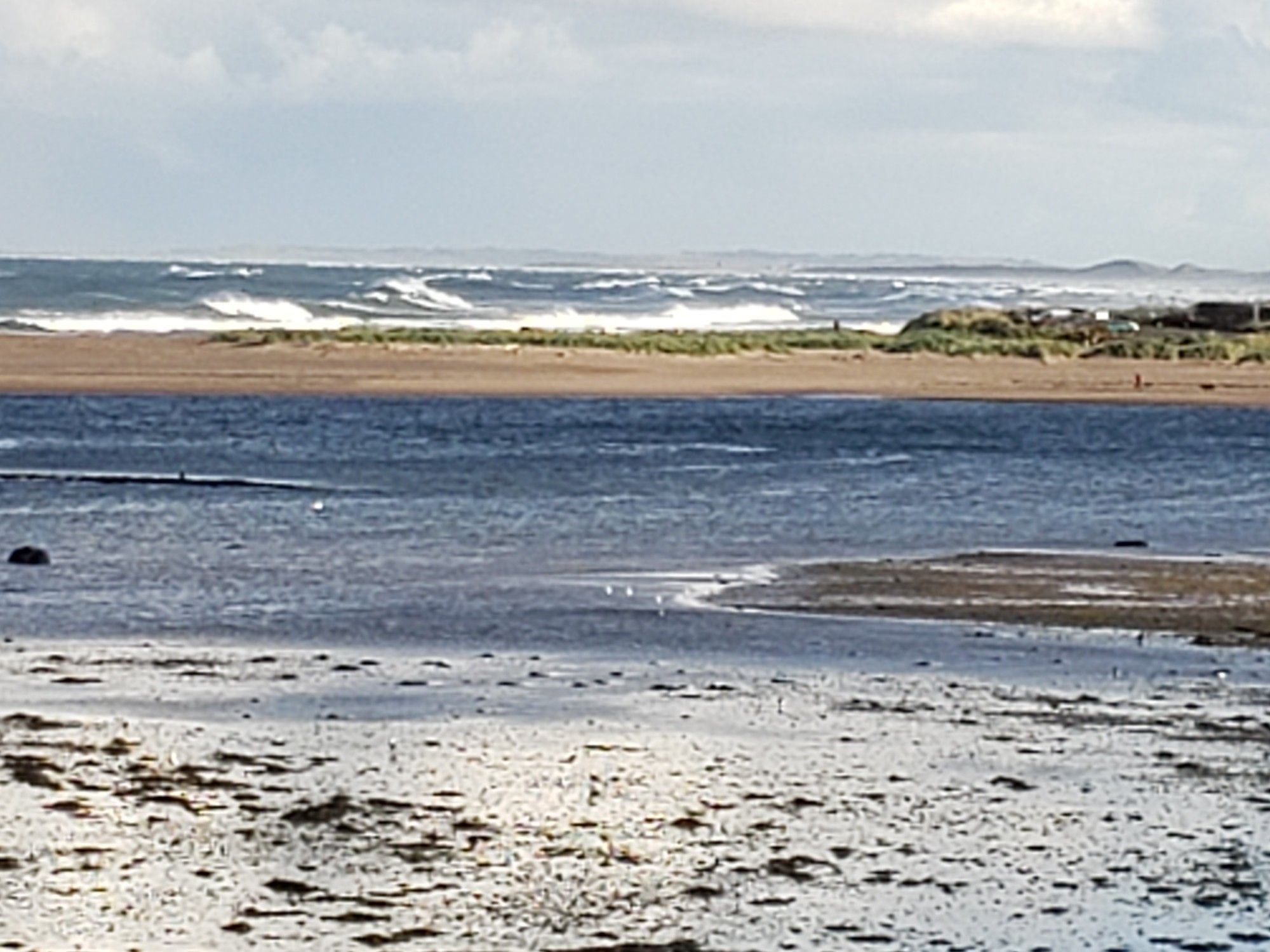 Low tide on a river estuary, with big waves crashing in deep blue sea beyond a sand bar