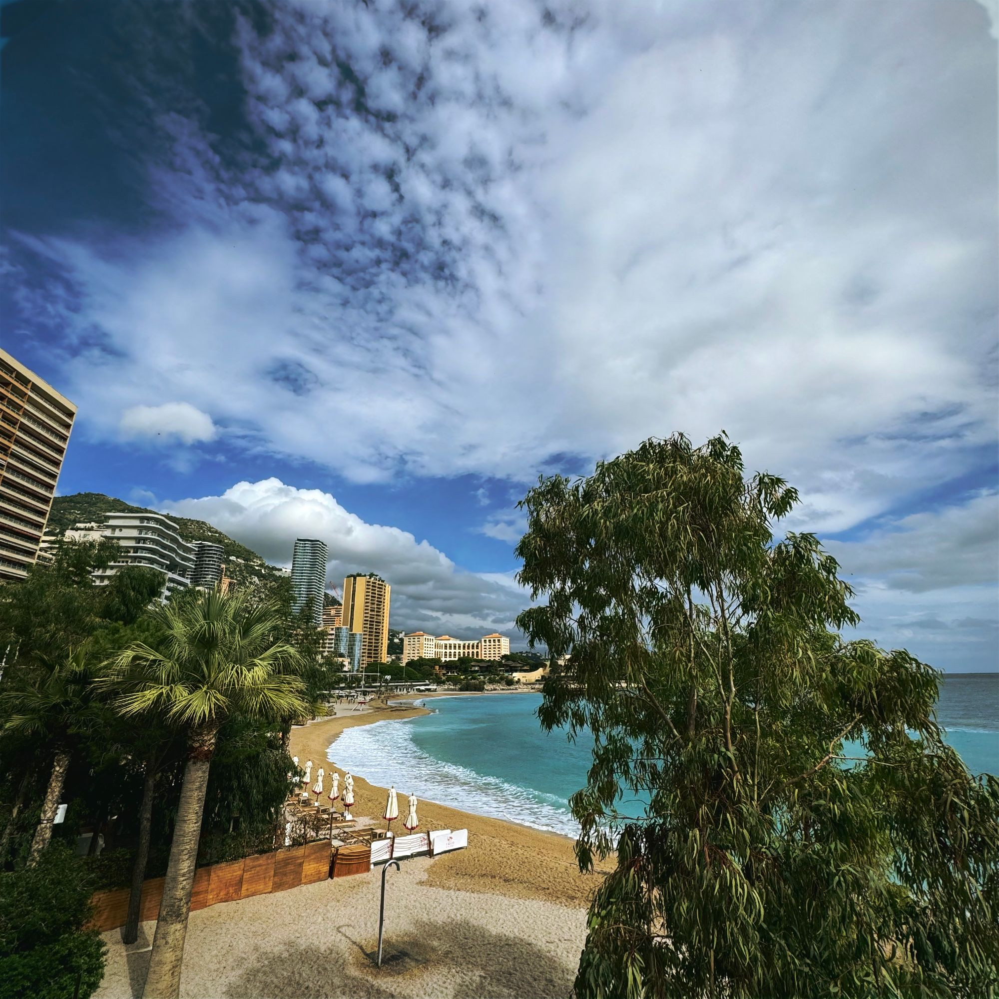 A view over Plage du Larvotto in Monaco, with a turquoise blue sea and golden sand, lined by trees. In the background, tall buildings, mountains, and mountainous clouds, under a deep blue sky full of fluffy cloud.