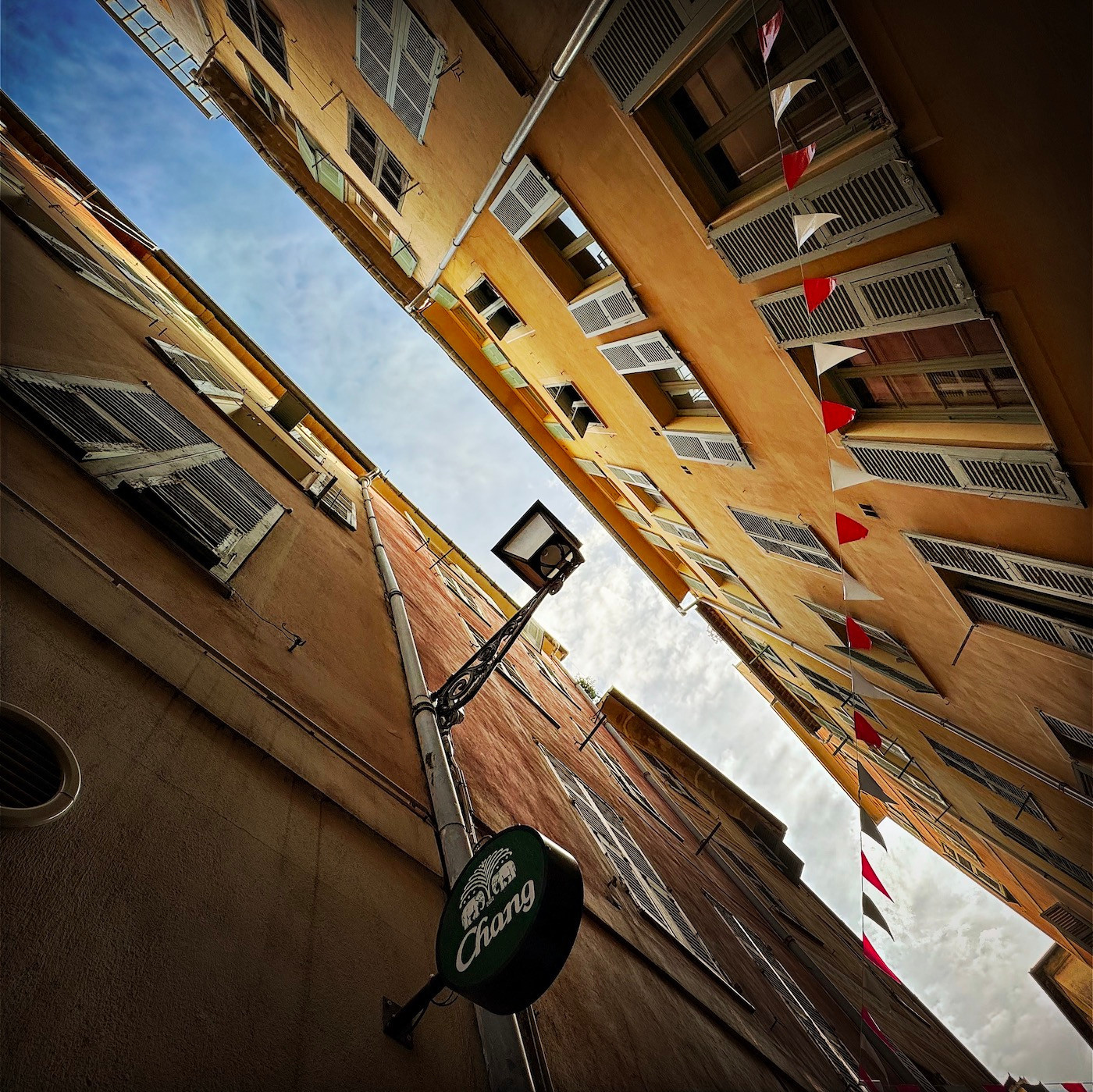 View upwards from a narrow street in Vieux Nice, between colourful painted walls, with a lamp post and a string of red and white flags. Above is a sky fading from rich blue to bubbly white cloud.