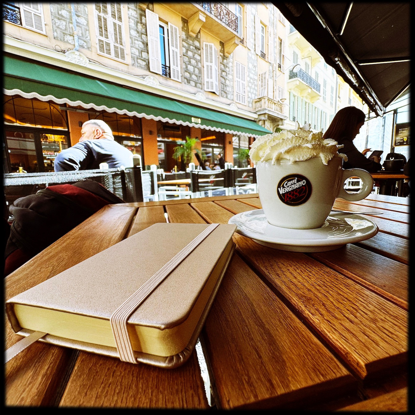 Poetry notebook and a Viennese cappucino with whipped cream, on a wooden table, looking out at a street lined with cafés and restaurants.