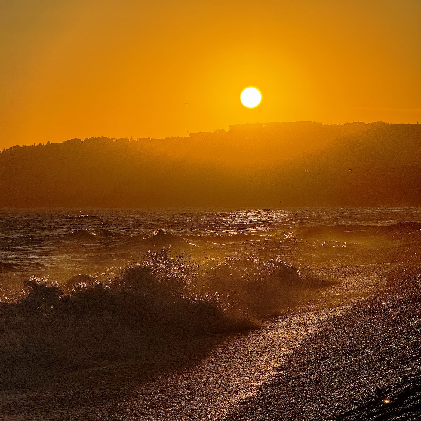 The setting sun painting the sky a rich orange, with the light reflecting off a shingle beach and the sea, as waves break on the shore and shatter into flying water drops.