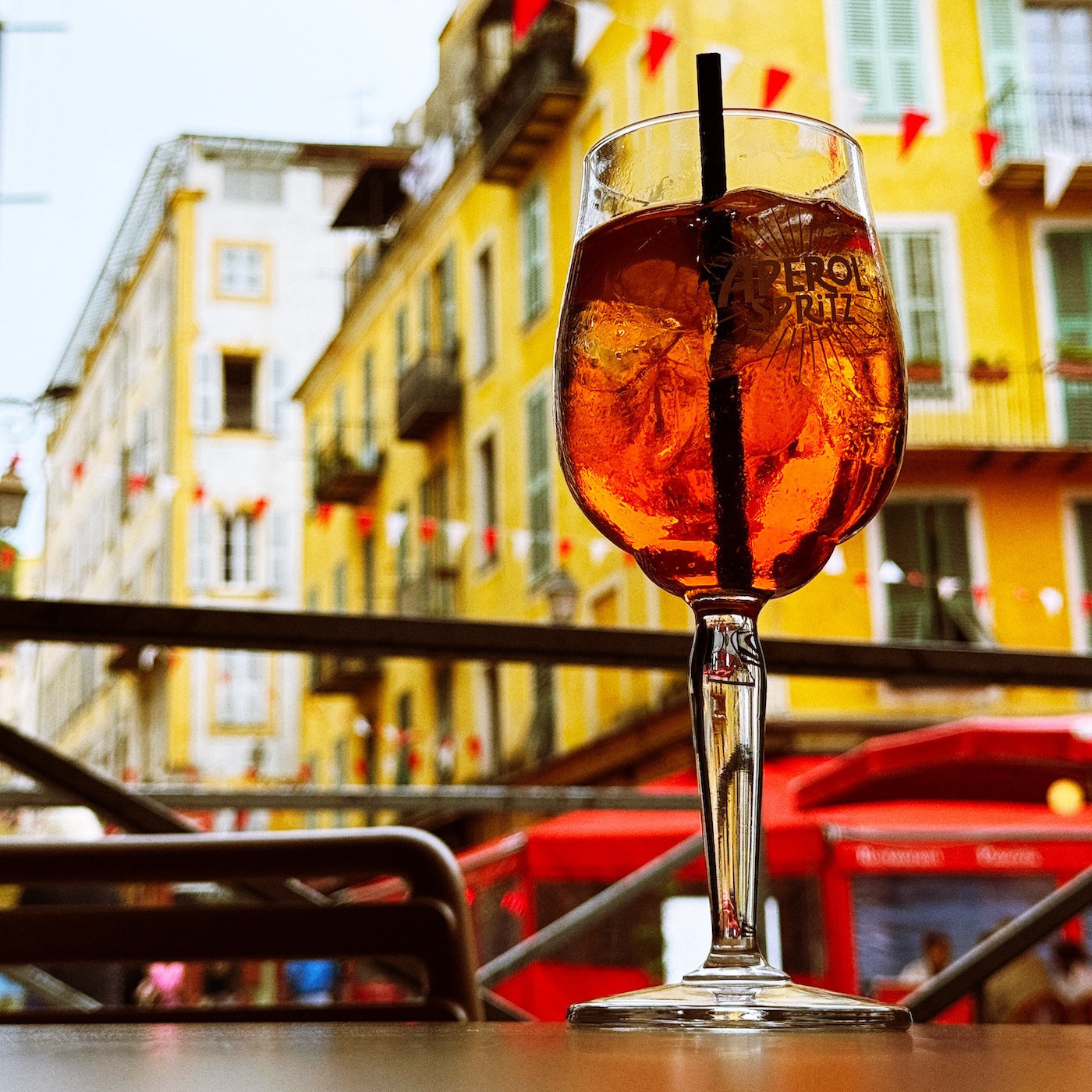 A bright orange Aperol Spritz on a table, in front of old yellow and white buildings covered with windows with wooden shutters.