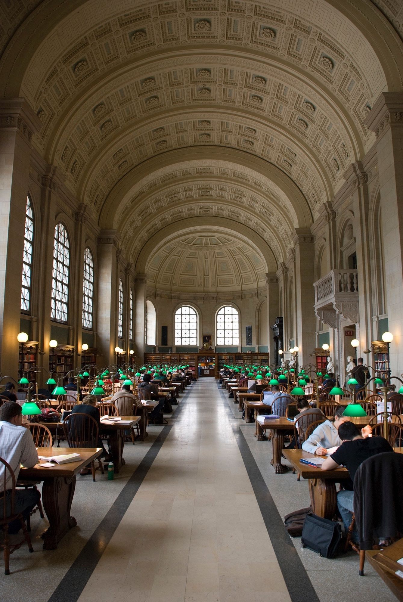 The reading room at the Boston Public Library. There's a long central corridor flanked by rows of desks with green-shaded reading lamps. A Arched windows run down one high wall on the left and continue around the far end of the room. A big vaulted ceiling above.