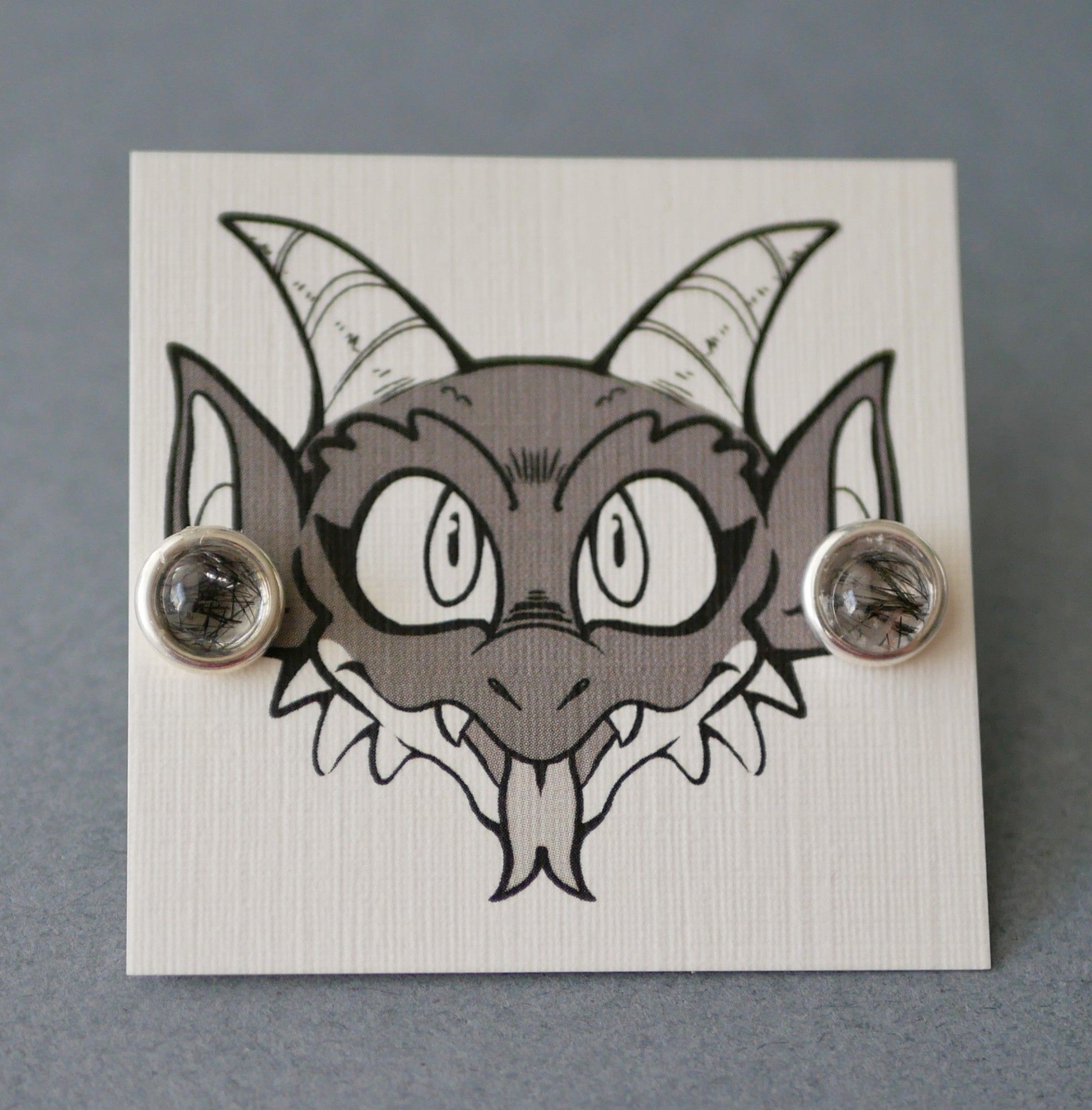 black tourmalinated earrings on an earring card with Bauble the kobold on it looking like he's wearing the earrings