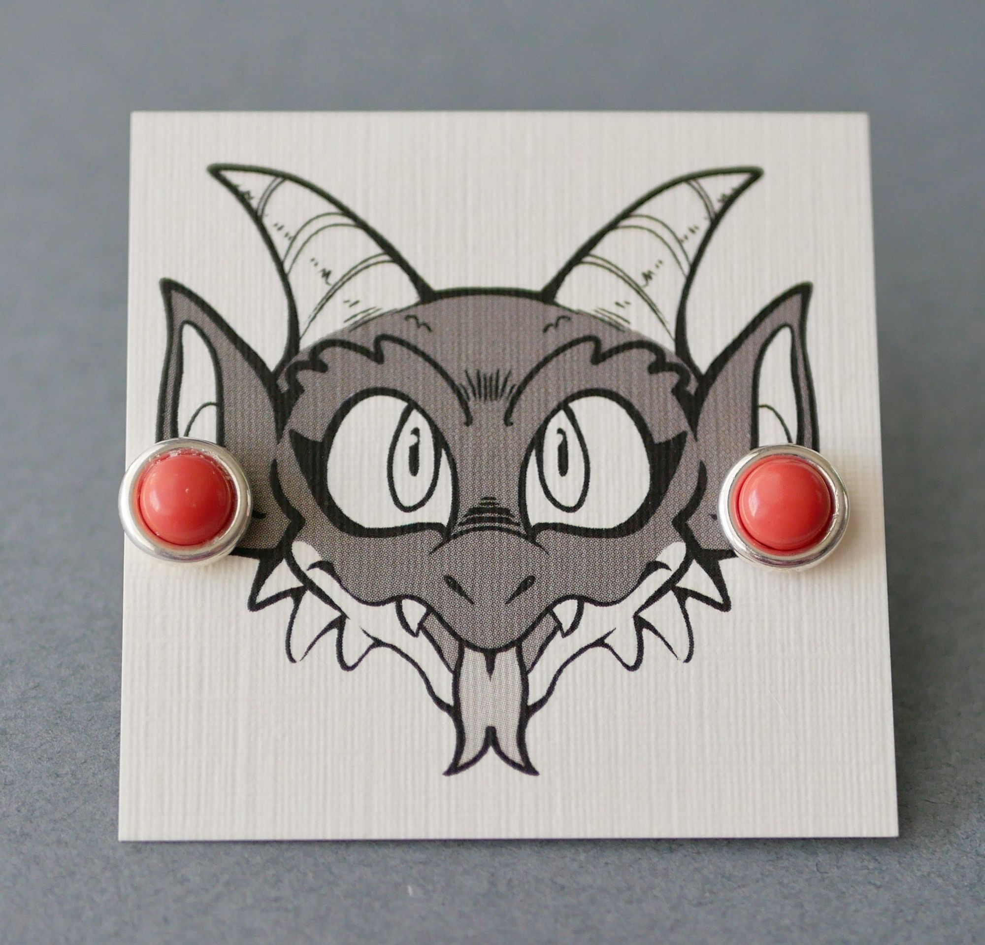 simulated salmon coral earrings on an earring card with Bauble the kobold on it looking like he's wearing the earrings