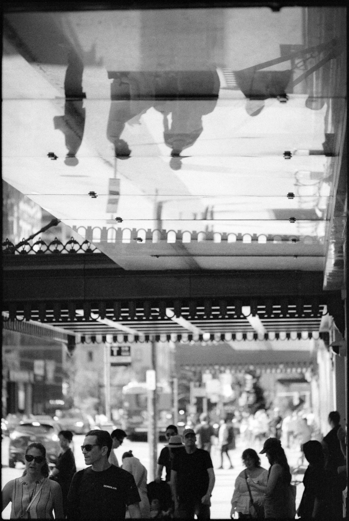 A crowd of people hide from the sun under a glass awning, the glass reflecting the street below.