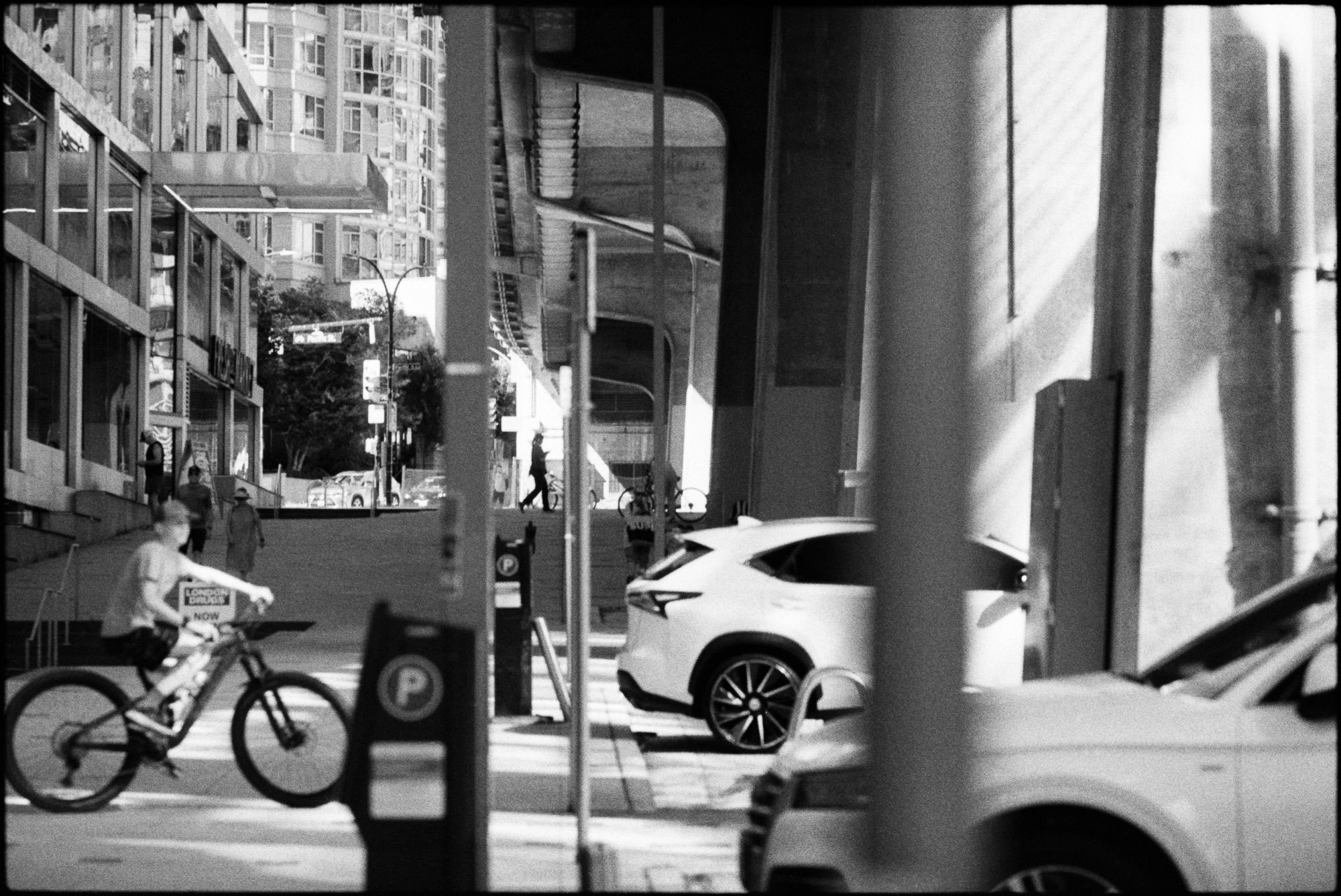 A child on a bike rides into the oppressive sun from under the cool shadows the Granville Street Bridge. Cars, pedestrians and cyclists are cast with slashes of light.