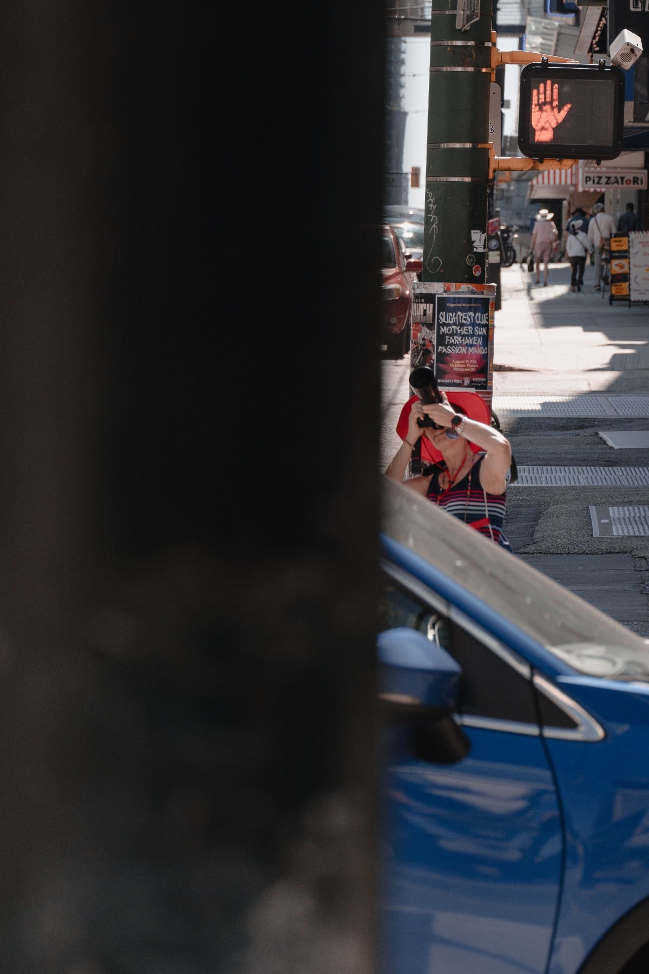 A photographer in red takes a shot of a downtown high-rise as a blue car passes before her.