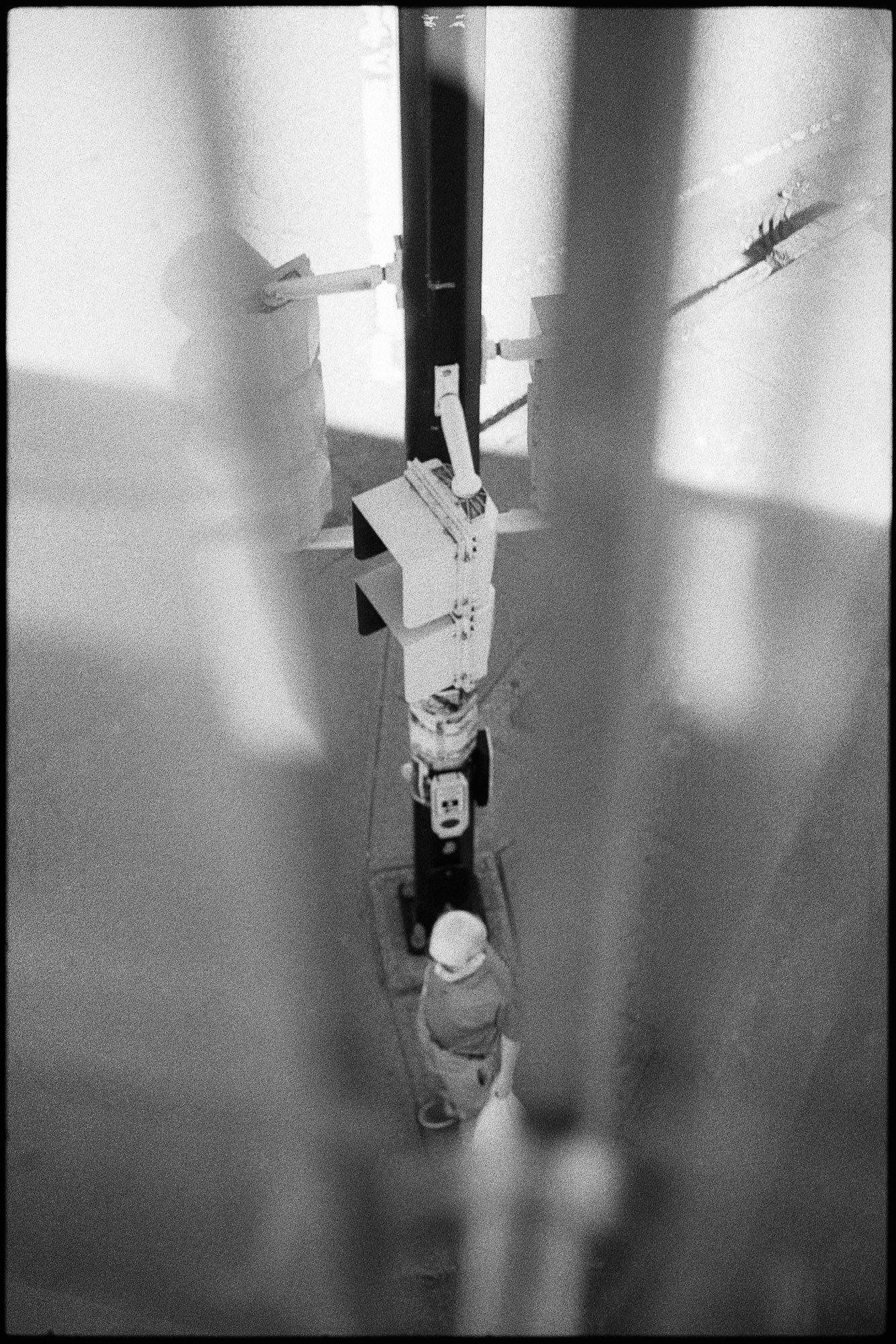 Black and photo of a bald man, viewed from above, waiting under an array of traffic signals to cross the road.