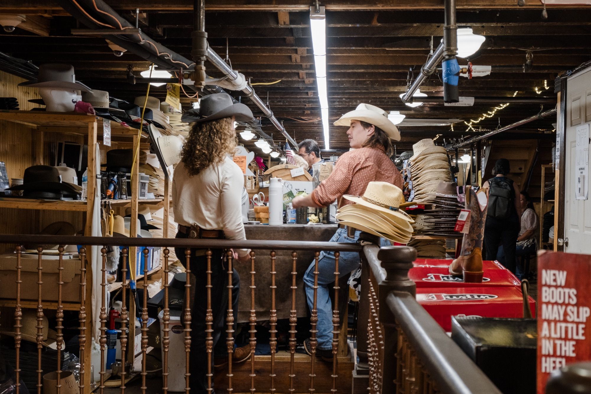 Two western ware employees converse amongst cowboy hats. A nearby sign says “New boots may slip a little in the heel.”