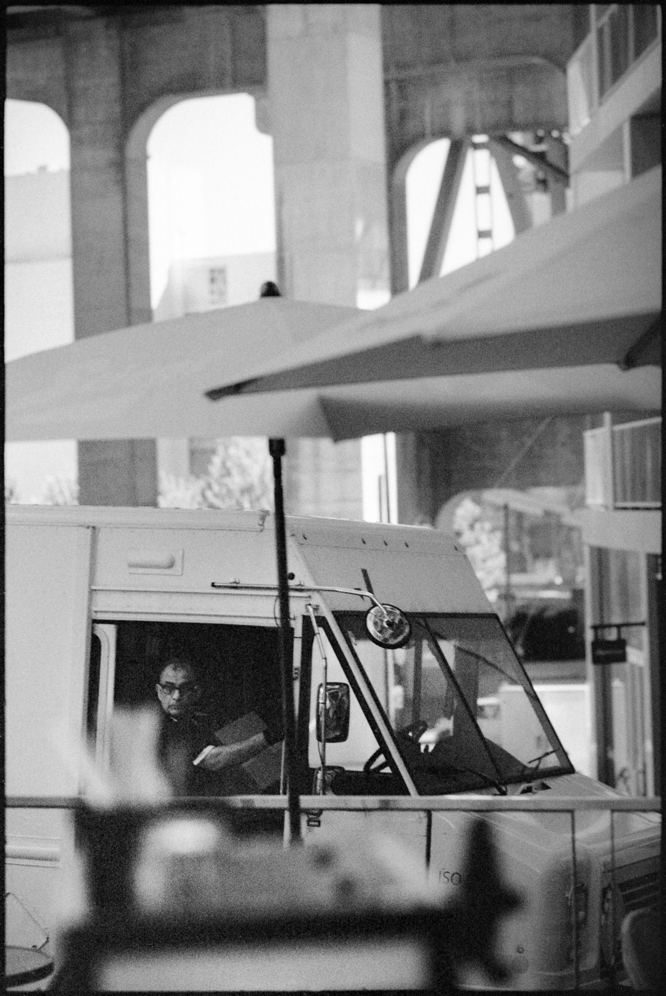 A courier leaves his van with a large package beneath his arm. Some umbrellas sit in the foreground and the structure of the Granville Street Bridge looms behind.