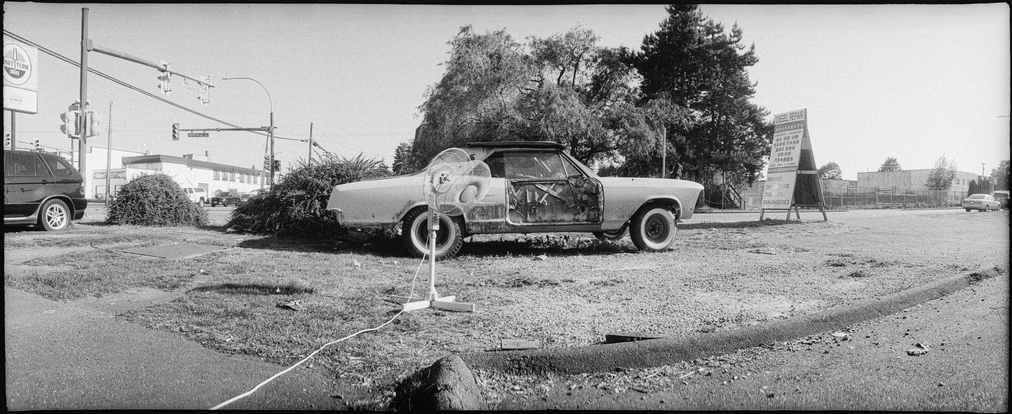 An old car missing a door panel sits behind a floor fan missing blades on a shabby industrial street.