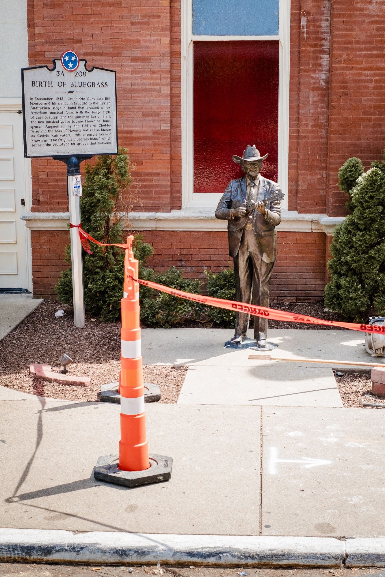 A statue of Bill Monroe with his mandolin stands amidst road and sidewalk construction and a plaque.