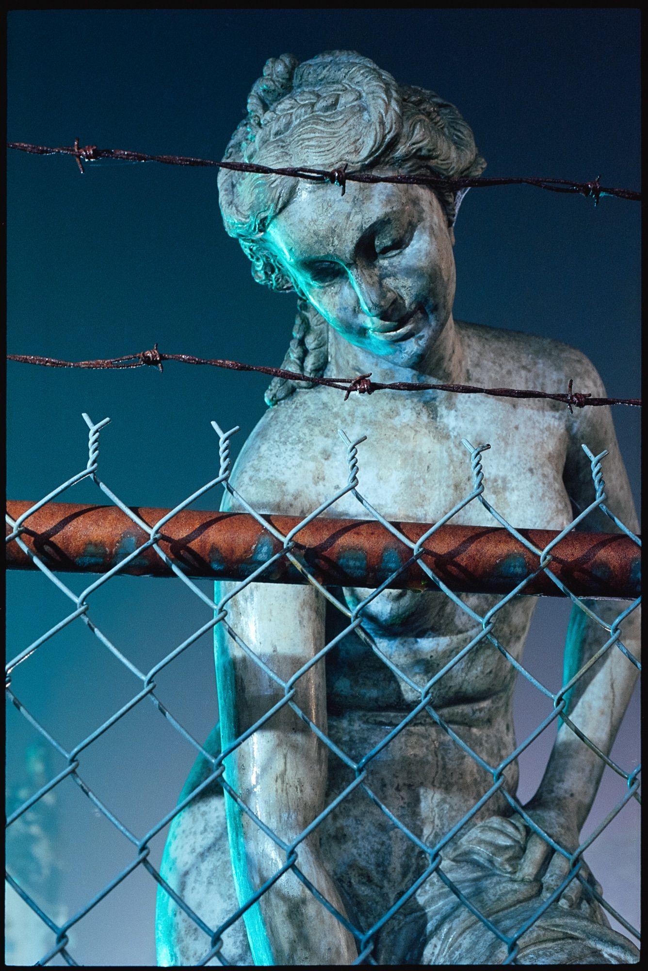 A lawn statue of a woman stares wistfully at the ground behind a barbed wire fence.