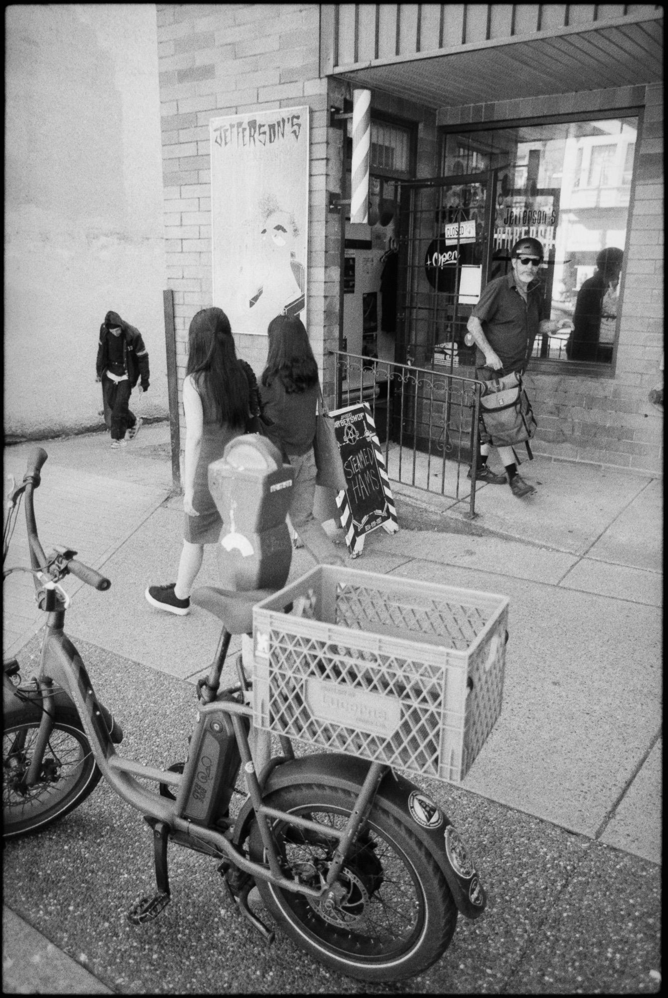 Black and white photo of pedestrians travelling in various directions in front of barber shop. A sandwich board reads “Steamed Hams.”