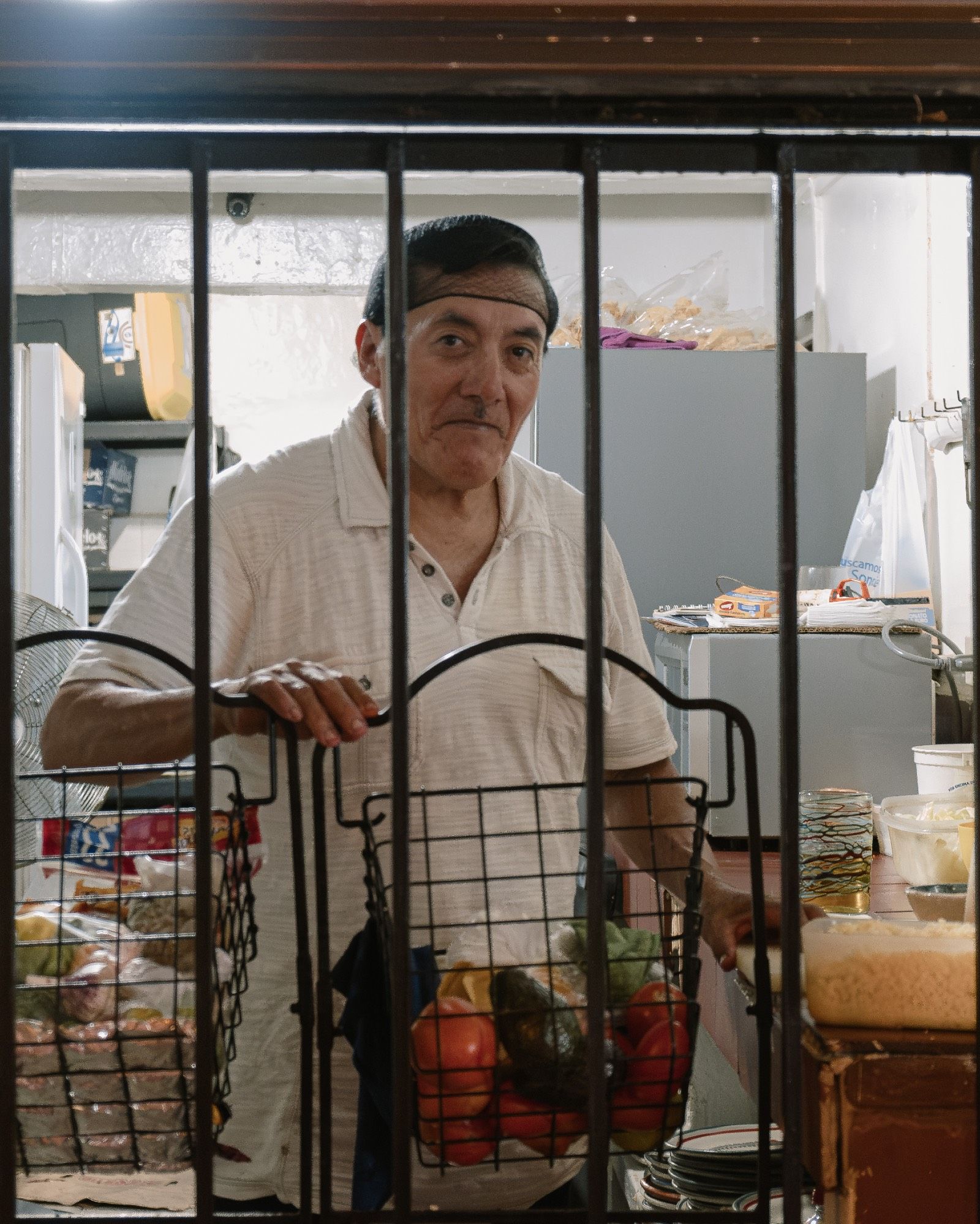 A chef in a hairnet looks through bars of his cluttered outdoor kitchen.