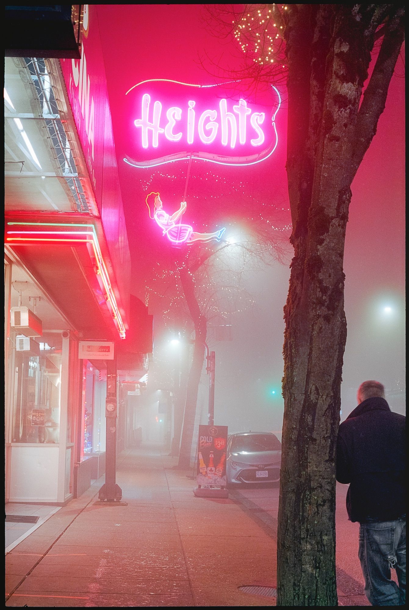 A man leans against a tree beneath a neon sign waiting for his ride to emerge from the glowing fog that envelops the evening.