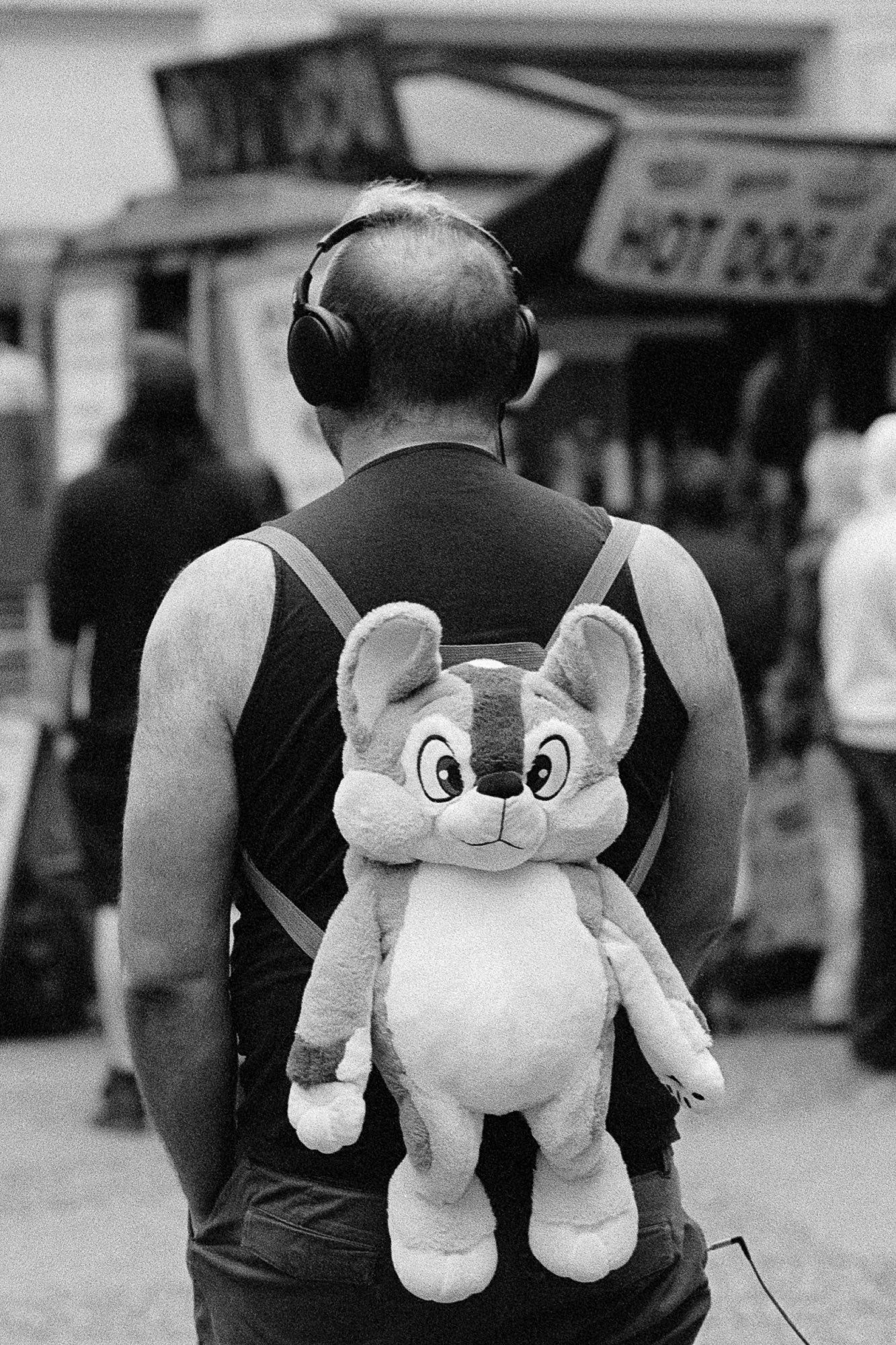 A man with headphones and a plush backpack plunges his hands into his pockets and watches the world pass by a hotdog stand.