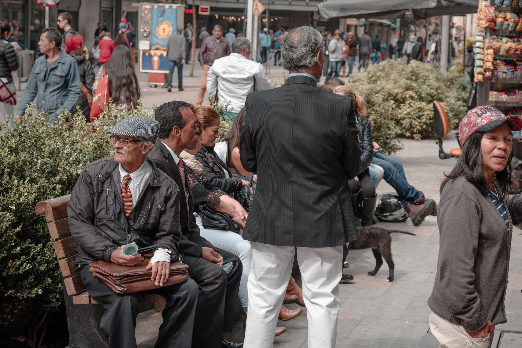 People rest on a bench. A man in the foreground looks past the horizon with a cup in one hand and a bandage on the other.