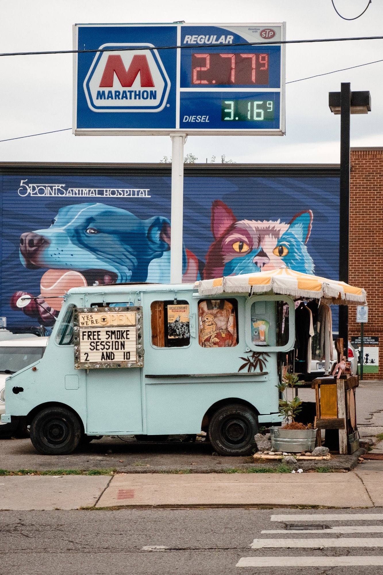 A smoke shop in a truck parks in a gas station lot with a mural of a cat and dog behind it.