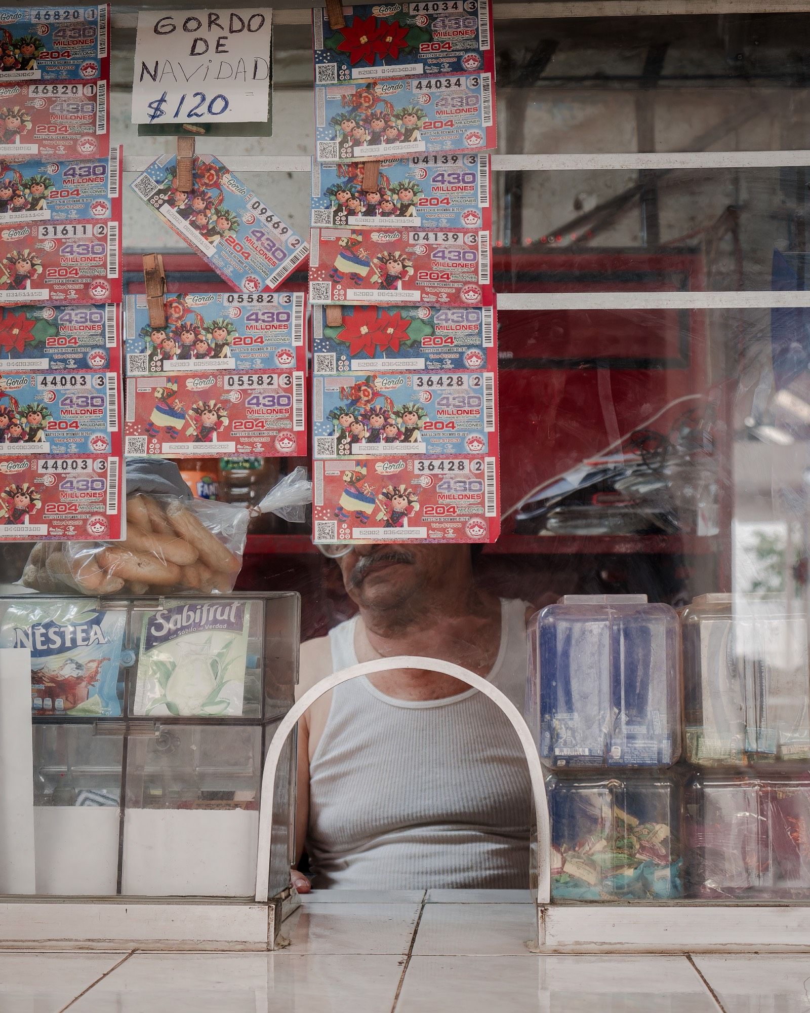 A man, face obscured by lottery tickets, sits behind a plexiglass window of his stall.