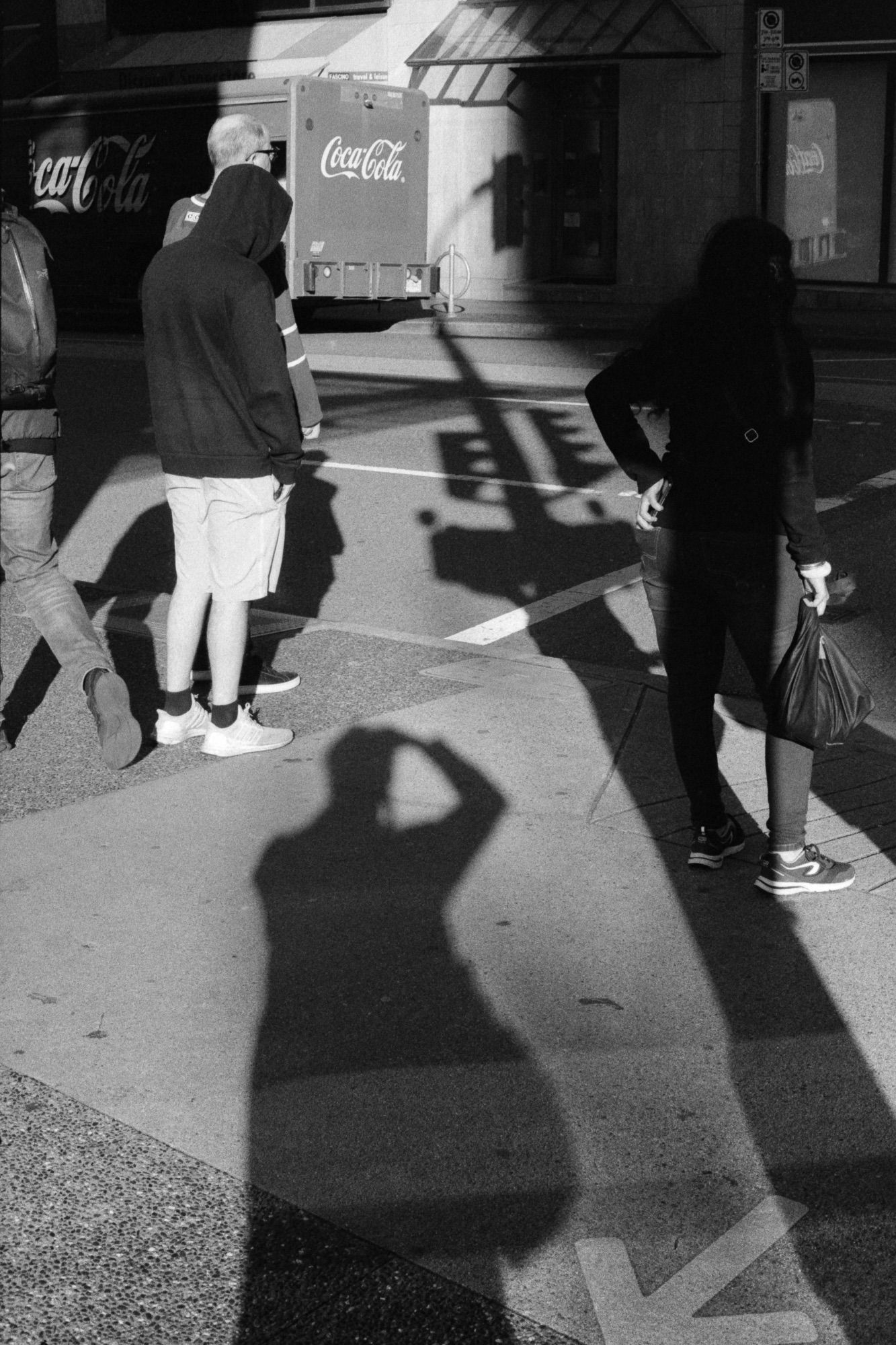 People waiting for the light to turn at an intersection amidst the long shadow of a light standard and the photographer.