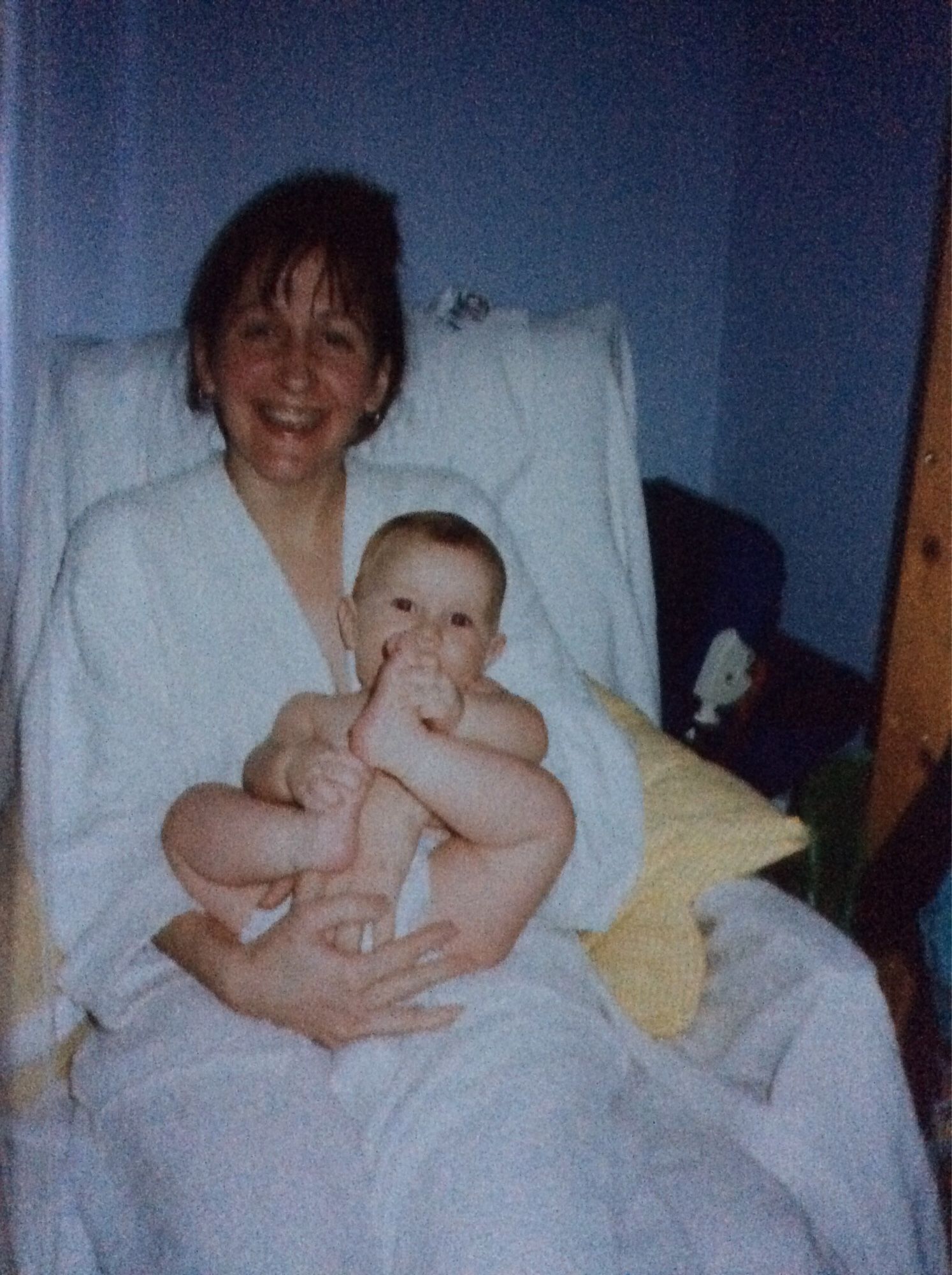 A mum sitting in a chair with her young son on her lap. The baby is wearing a nappy and holding one foot up to his mouth. They both look very happy ❤️