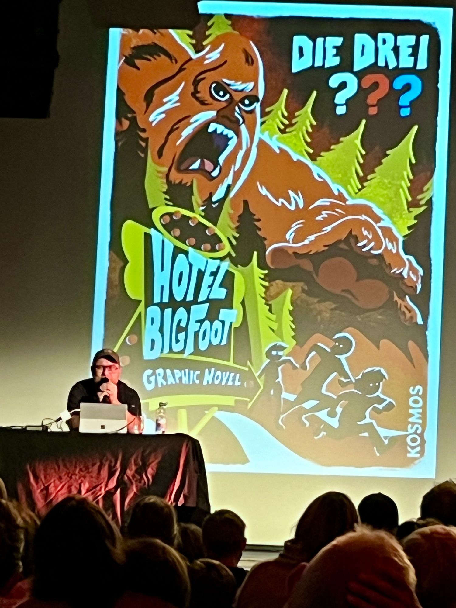 Ein Autor spricht bei einer Präsentation, mit einer großen Projektion hinter sich, die das Cover einer Graphic Novel mit dem Titel "Hotel Bigfoot" zeigt. Das Cover zeigt einen wütenden Bigfoot, umgeben von Bäumen, zusammen mit Silhouetten von laufenden Charakteren.