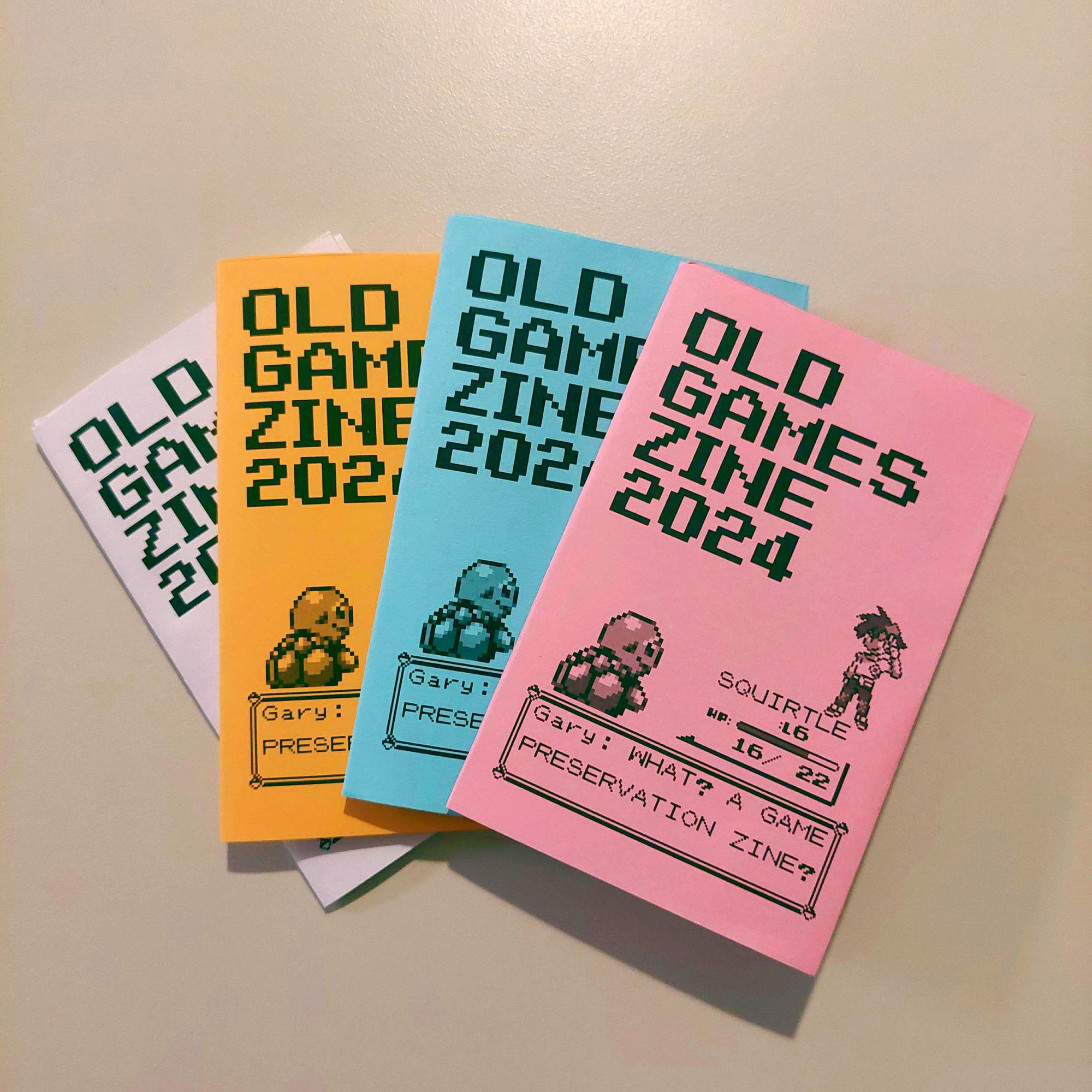 A collection of "Old Game Zine 2024" prints in different colours. On the front, a pokmeon trainer named gary yells "WHAT? A GAME PRESERVATION ZINE?"