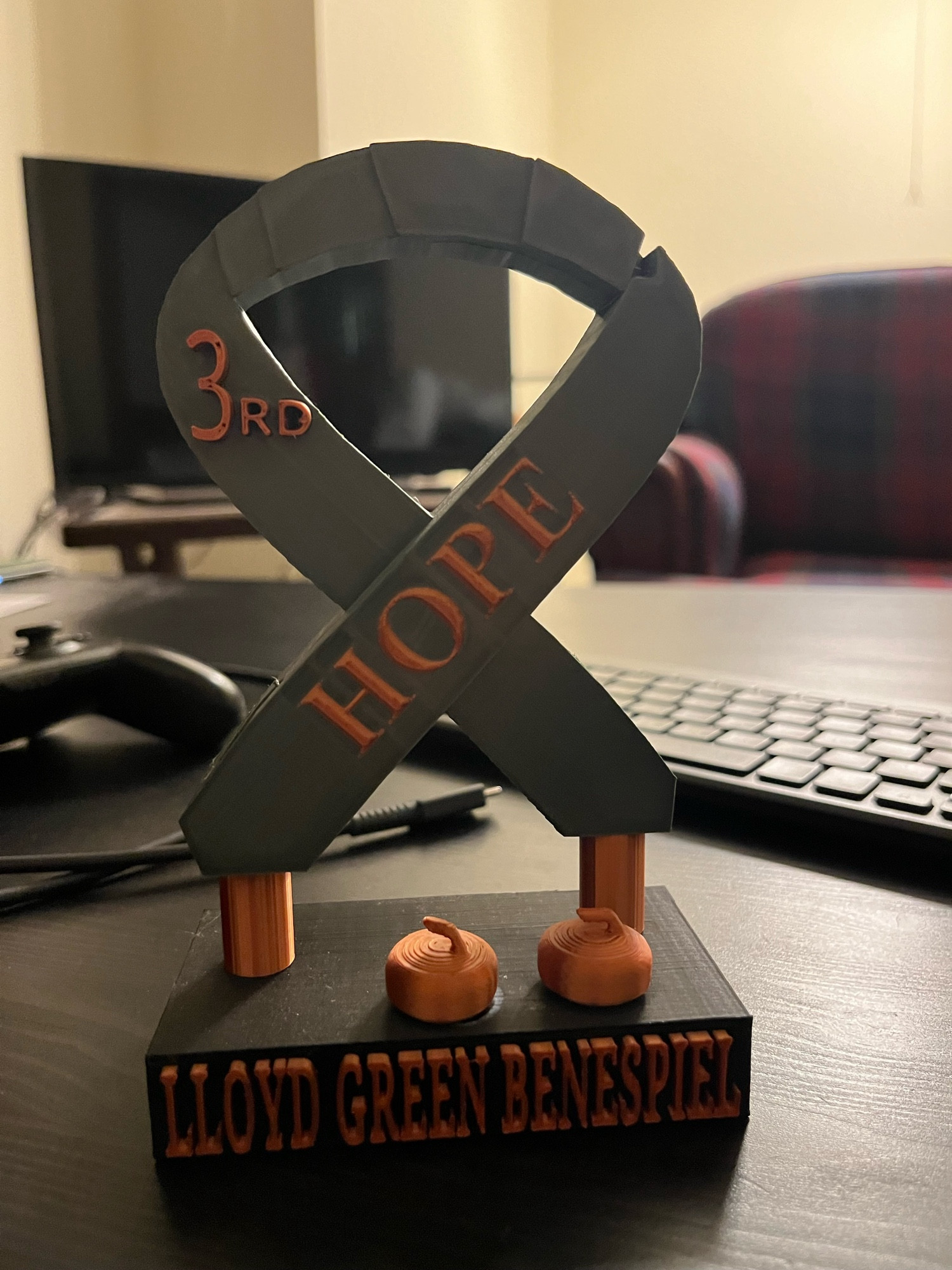 A trophy in the shape of a green cancer awareness ribbon on a base labeled “Lloyd Green Benespiel.” The ribbon says “HOPE” and “3rd.” There are two curling stones on the base in front of the ribbon. All of the accents are in a bronze color.