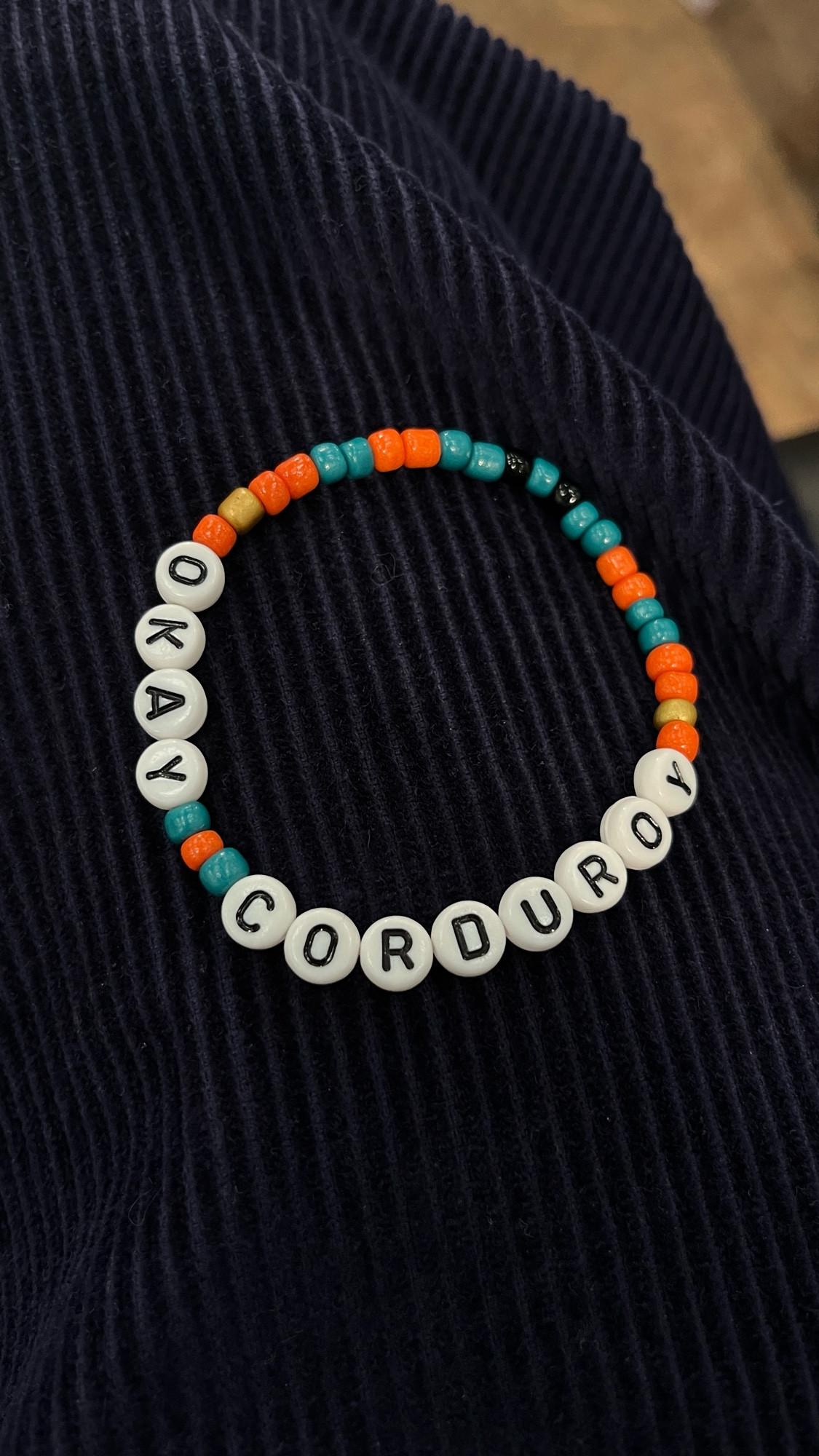 A handmade bracelet reading ‘okay courdory’ in the colours of the Worlds Beyond Number podcast on a pair of navy corduroy trousers