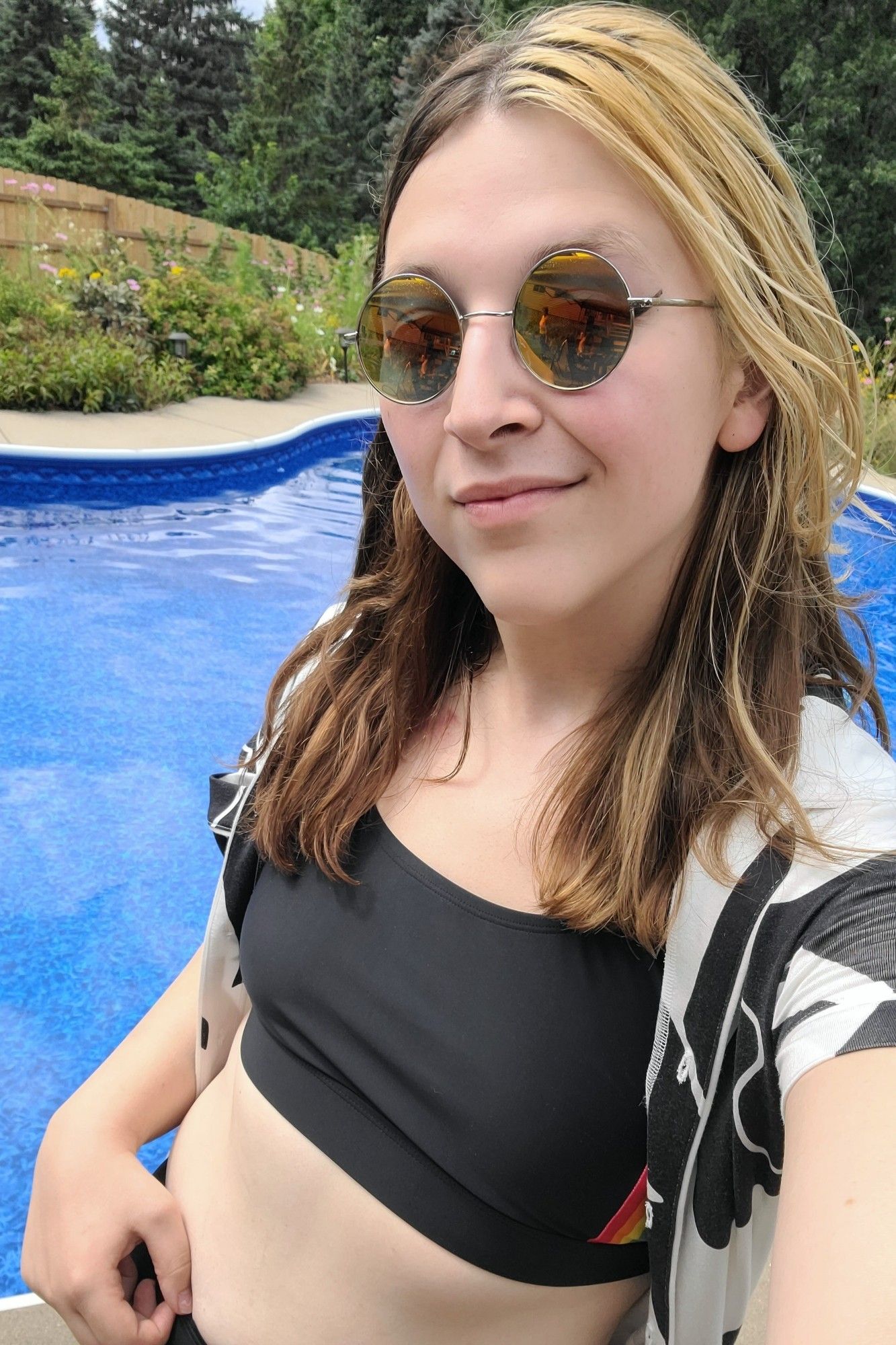 picture of me, a white trans woman, wearing a black swimsuit and a black and white patterned button up, standing in front of a pool