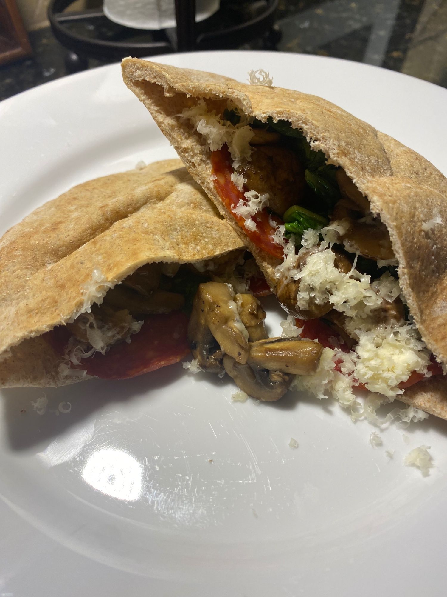 A white plate holds two halves of a split whole wheat pita, each stuffed with two thin slices of Spanish-style chorizo, sautéed turnip greens (from CSA box), sliced cremini mushrooms sautéed with butter, EVOO and a splash of sherry, and grated Bellavitano chili lime cheese.