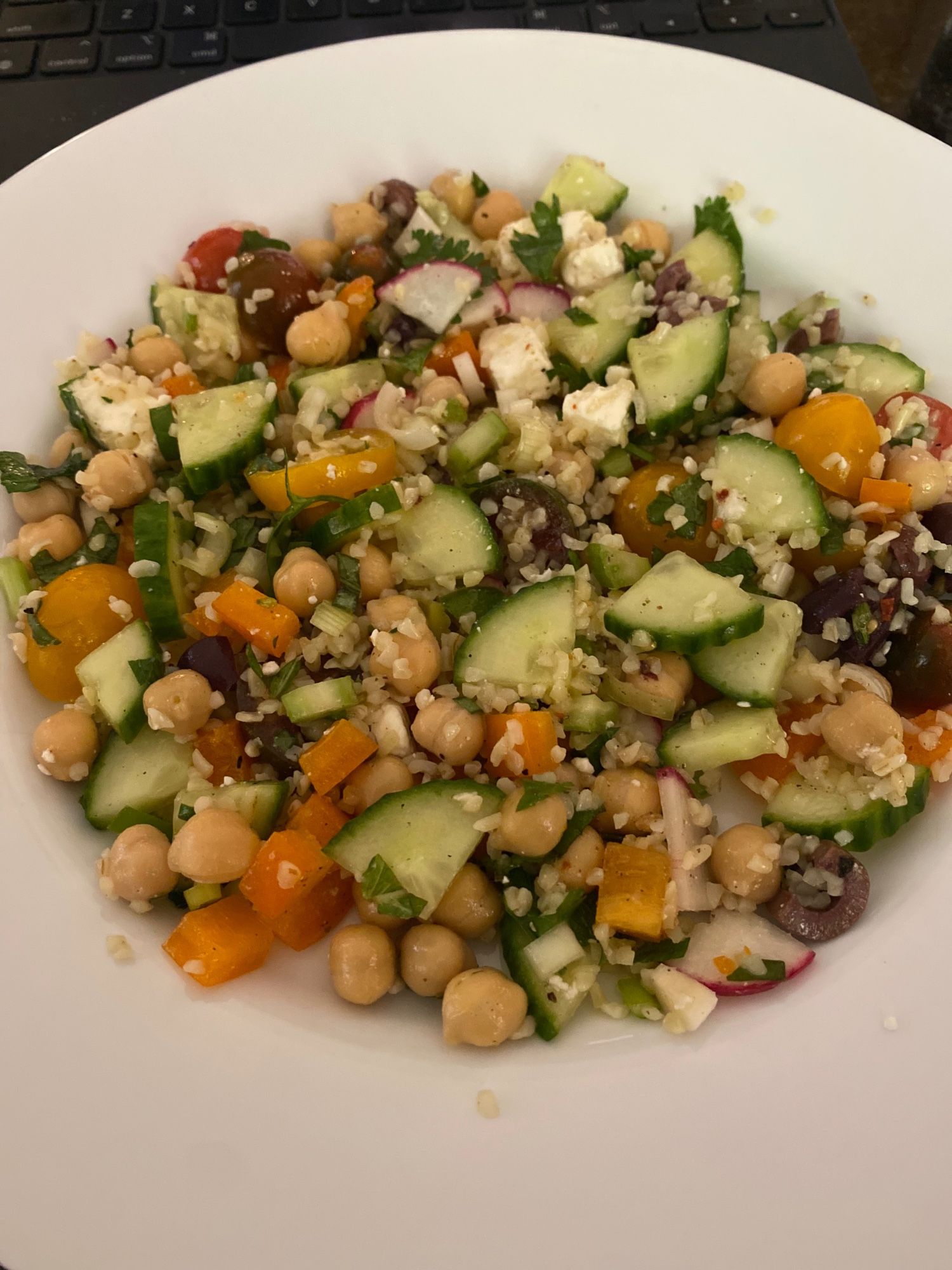 White plate with colorful mixture of vegetables and other ingredients. Mixture includes (all diced or chopped): yellow pepper, jalapeño, radishes, celery, grape tomatoes, green onions, chickpeas, Kalamata olives, capers, bulgur, feta, fresh basil, parsley and cilantro, olive oil, Aleppo pepper, Za’atar, salt and pepper.