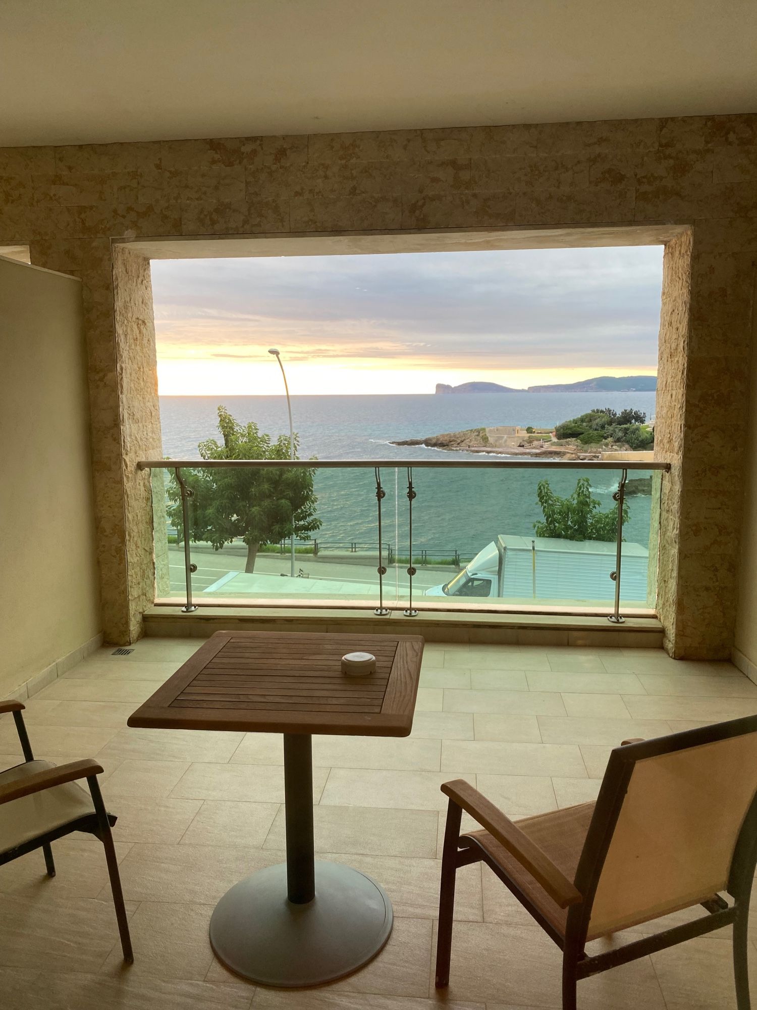 A view from a hotel room of a small patio with two chairs and a wooden table in front of a framed view of the sea with a setting sunk, distant cliffs, trees and rock outcroppings.