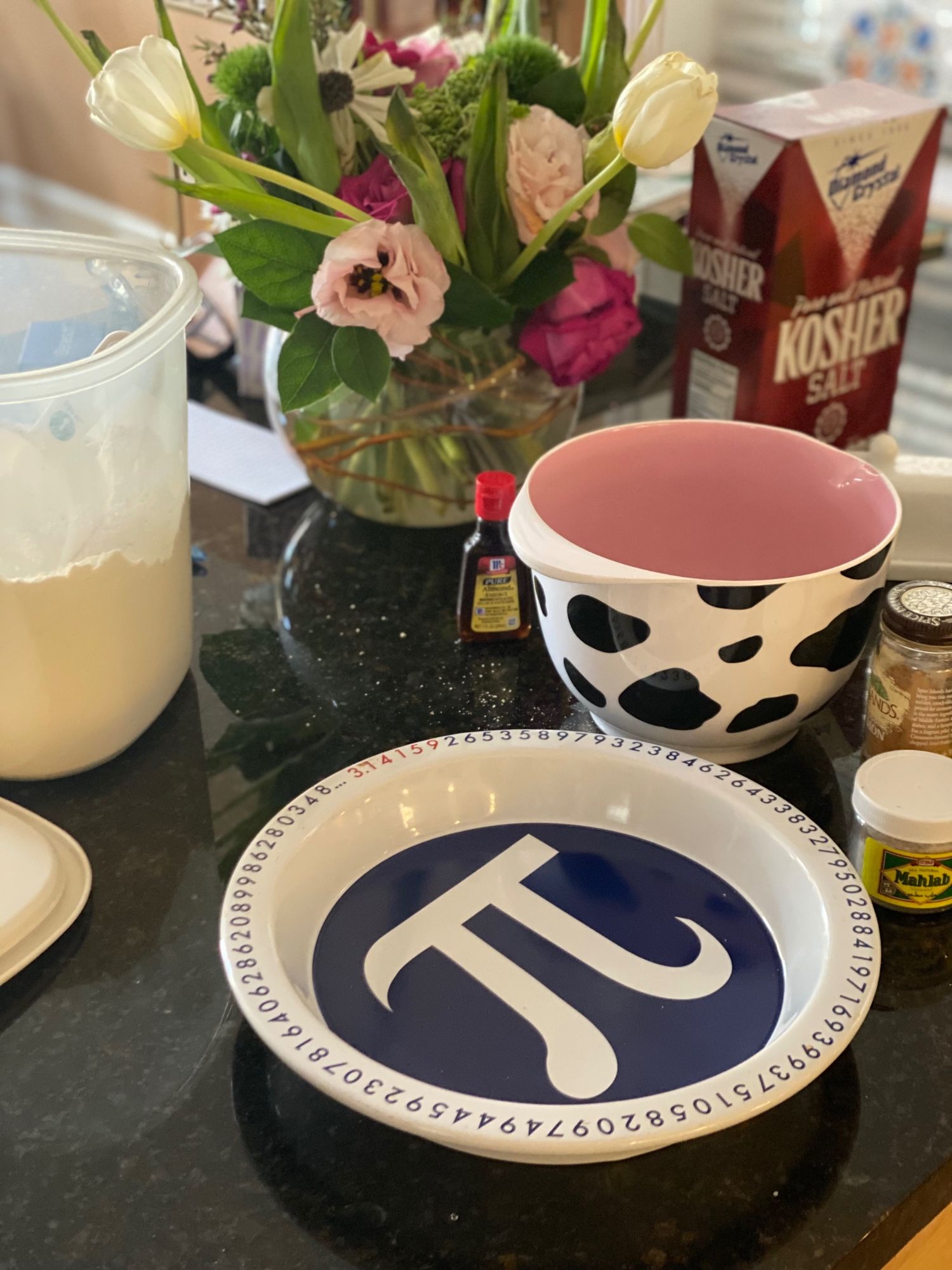 A white and blue pie plate with the Greek letter, Pi, printed inside and rimmed with the numerals that comprise mathematical pi. A back granite countertop holds the pie plate, an open tub of flour, a vase full of flowers, a small bottle of almond extract, a cow-print bowl with a pink inside, a box of kosher salt, a small jar of cinnamon and a small container of mahlab.