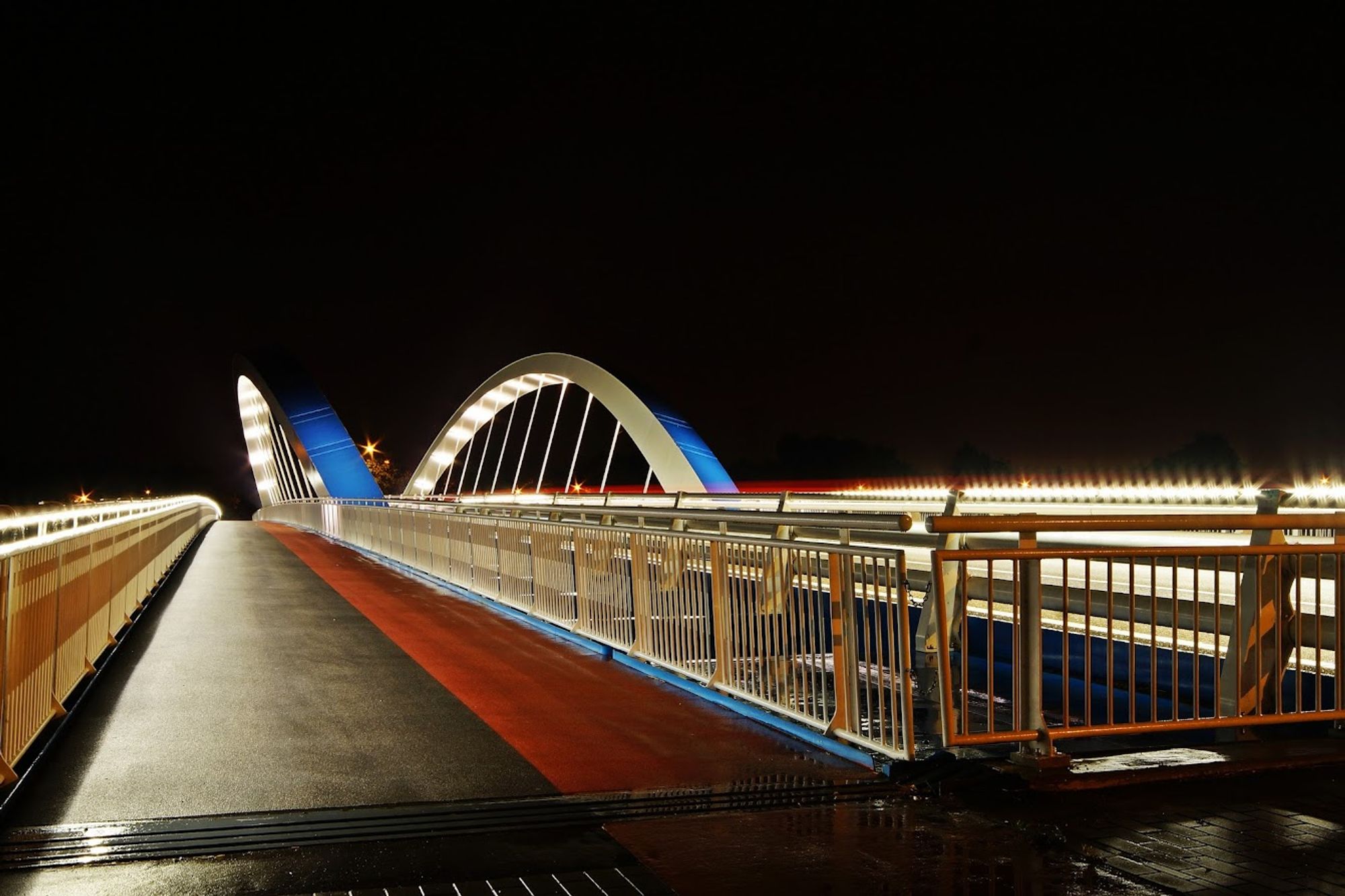 Blick auf den Gehweg und Radstreifen einer  beleuchteten Brücke in der Nacht. Die äußeren Geländer links und rechts sind beleuchtet, Ebenso die beiden Brückenbögen die in blau weiß gehalten sind.