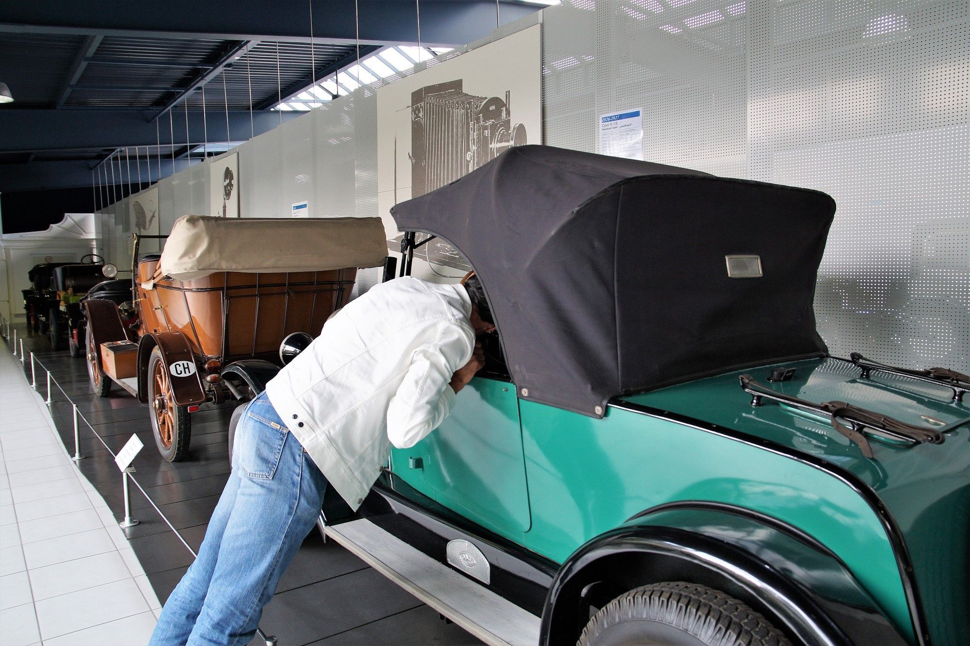 Ein Mann in blauer Jeans und weißer Jeansjacke beugt sich weit vor und schaut in einen grünen Oldtimer mit schwarzem Stoffdach. Er stützt sich an der Fahrertür ab. Sein Kopf ist im Auto.