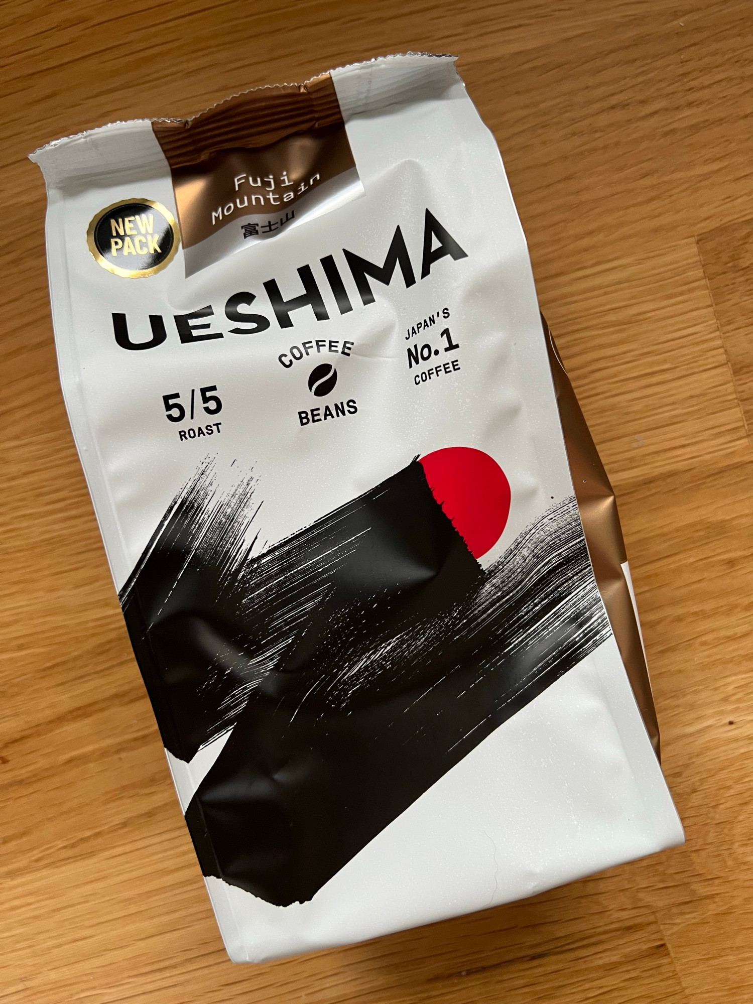 A pack of Ueshima brand coffee beans resting on an oak work surface. The pack is white with bronze coloured sides and features a wide black zigzag brush stroke partially obscuring a red circle. Above this are details of the roast strength and above that the brand name in capitals in a medium weight sans serif typeface.