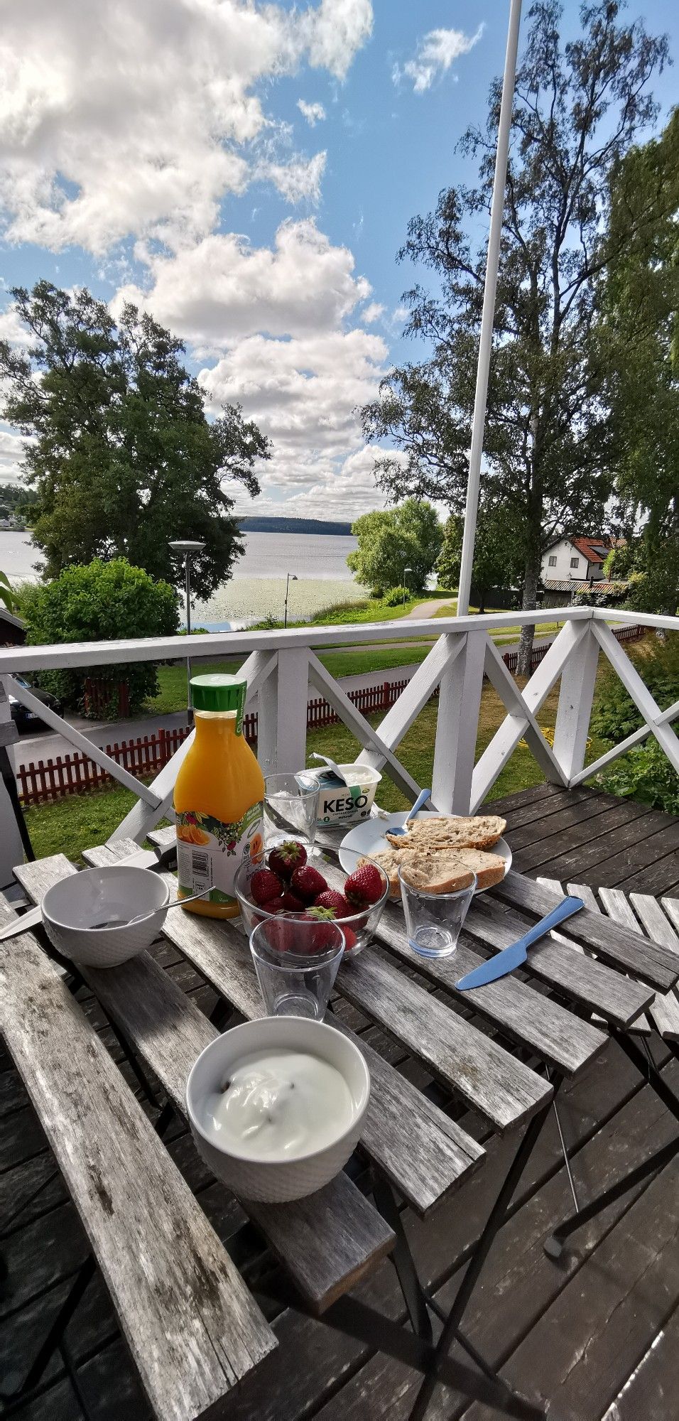 Photo d'une petite table de terrasse faite de planches en bois, avec un petit-déjeuner posé dessus (café, bouteille de jus d'orange, fraises dans un bol en verre, yoghourt dans un bol blanc, tranches de pain, verres vides et couteaux)
Derrière la barrière blanche de la terrasse, la vue donne sur un lac et des arbres. Le ciel est bleu avec des nuages blancs. 