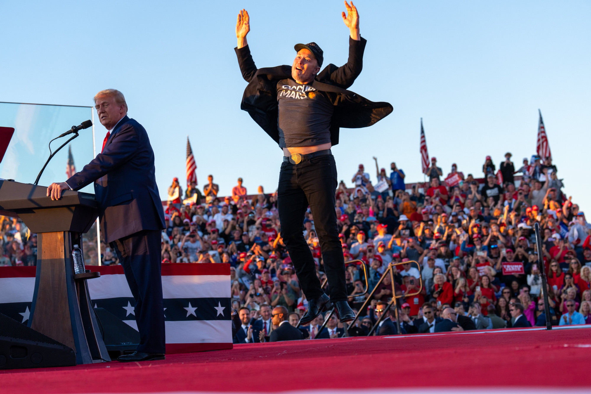 Donald Trump introduces Elon Musk during a rally in Butler, Pa. Musk is captured jumping in the most awkward way possible, with his hands above his head and his shirt lifting up to show some of his stomach