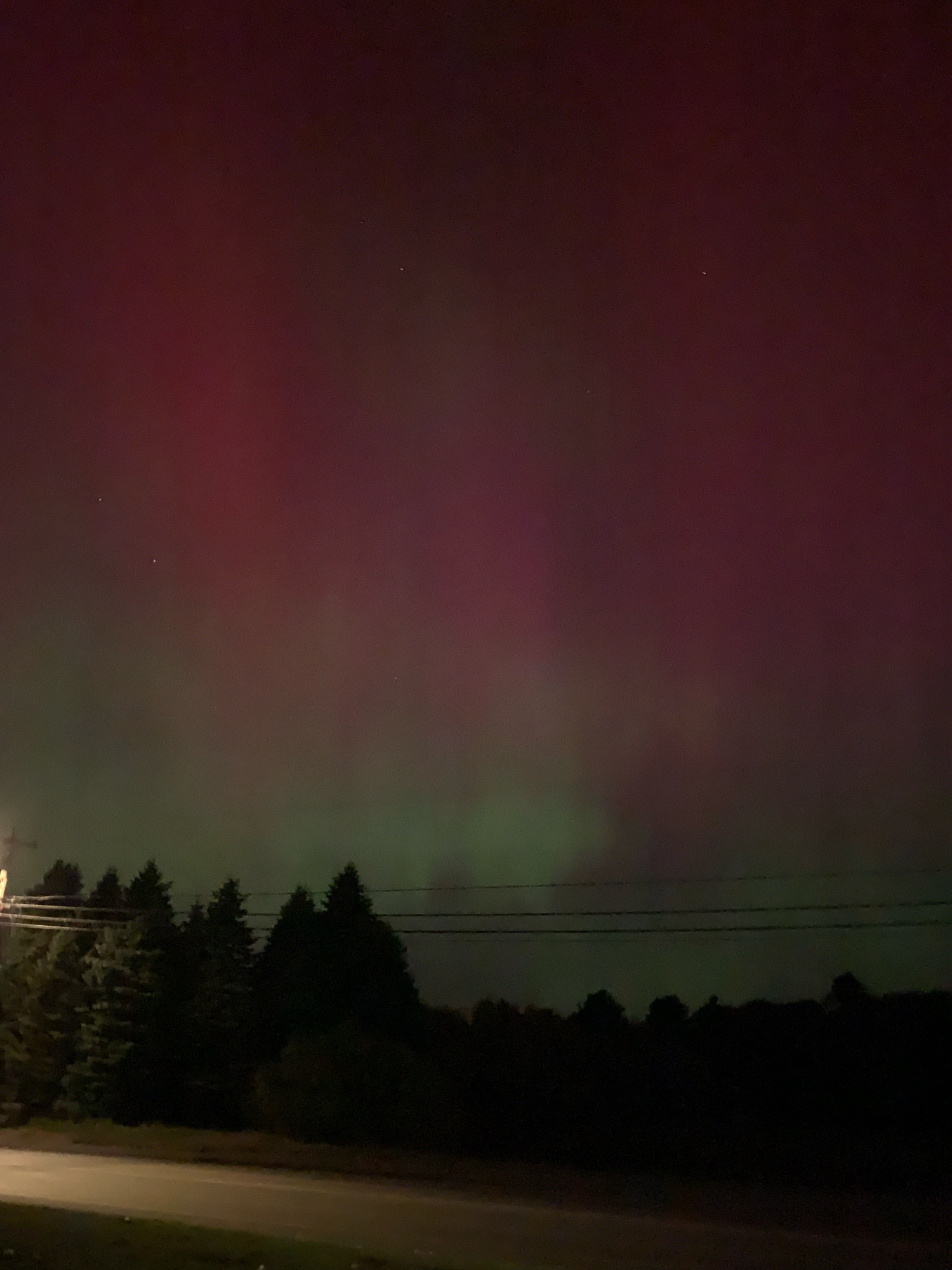 Photo of red and green aurora in the sky above a partially lit road and trees