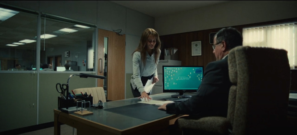 Ella Purnell playing Rhiannon in tv show, Sweetpea, she is handing her boss an application for a job role, in the background is his PCs desktop screen with icons all messed up. 