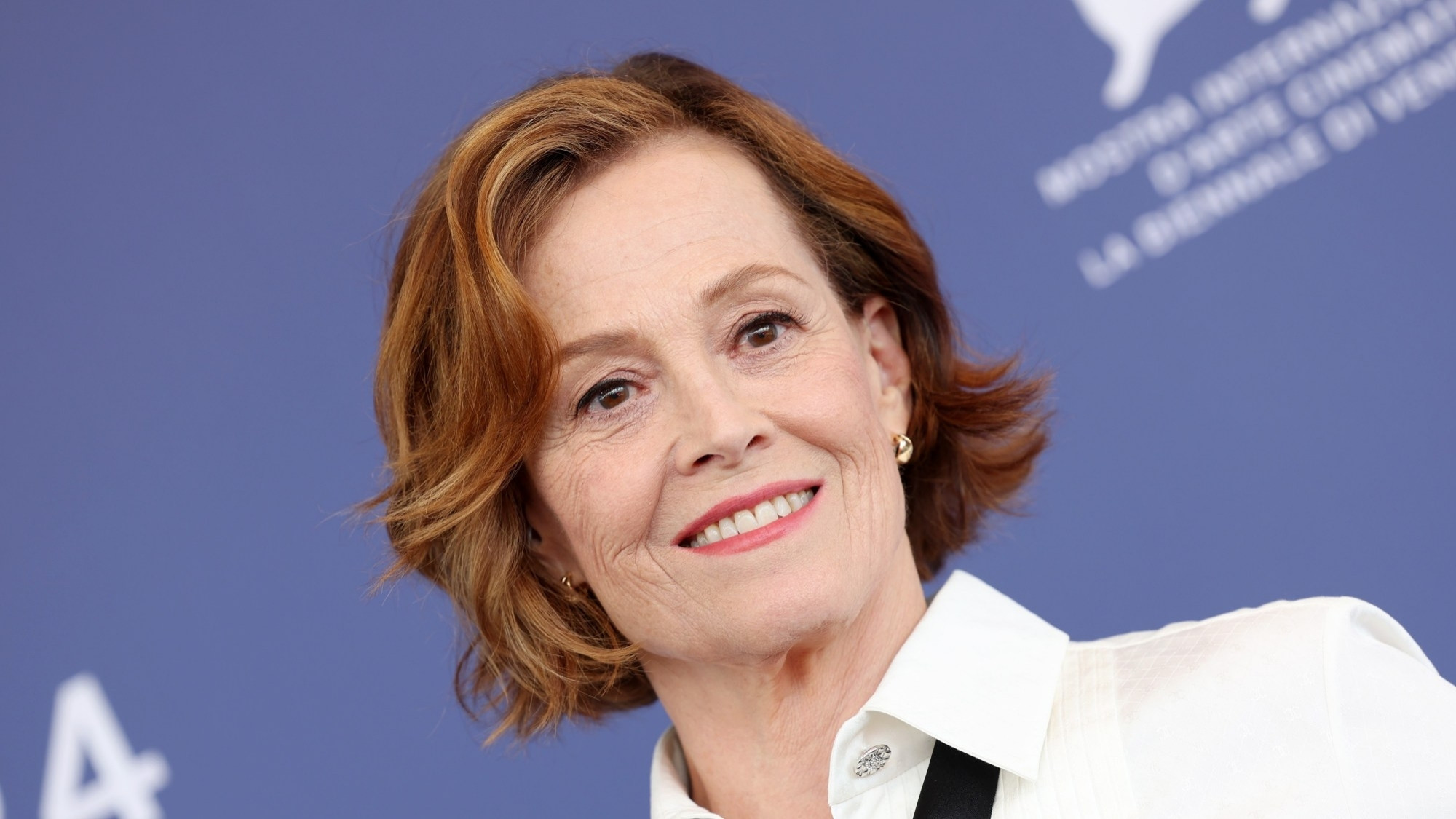 A photo of Sigourney Weaver in white shirt and black tie, and chin length wavy hair.