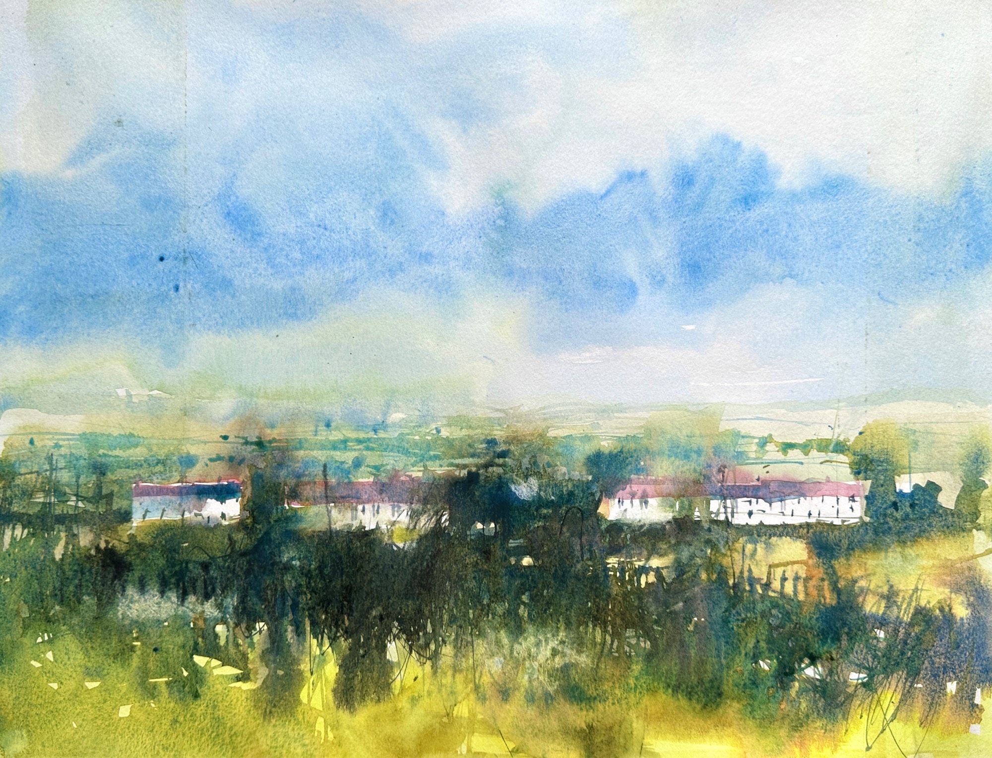 A view across winter fields and hedges to 3 rows of reddish roofed houses on the outskirts of the village. A blue/green distant background of fields and trees all under a cloudy blue sky