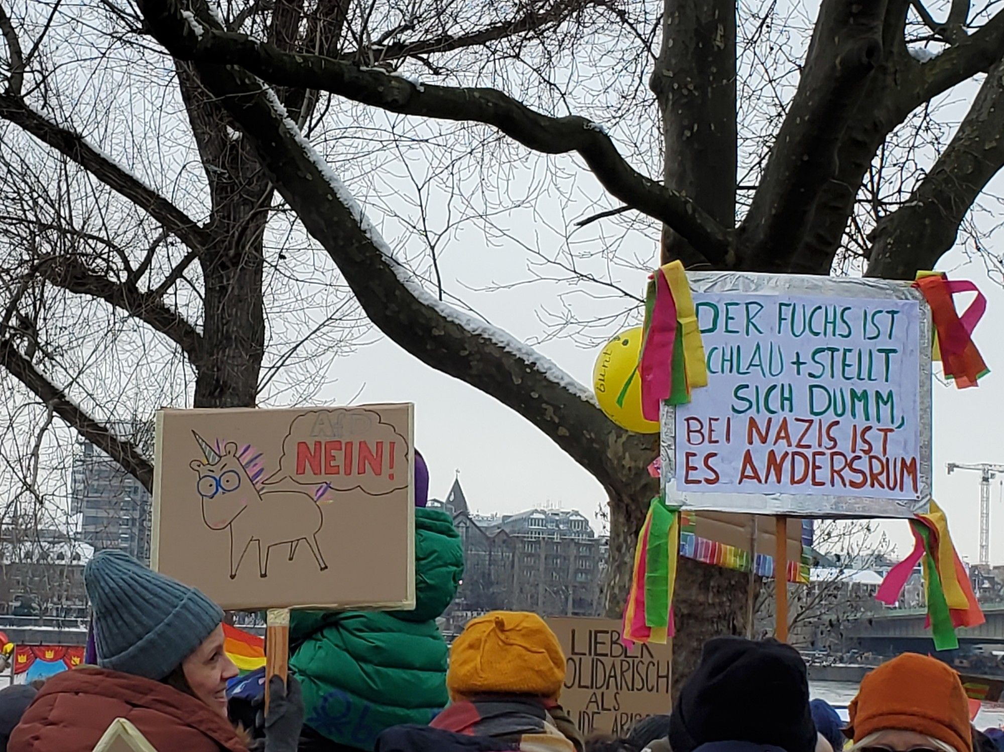 Two signs here: one, a "nein-horn" (a unicorn with a speech bubble, saying "AfD NEIN!"), and one festooned with multicolored ribbons which reads "Der Fuchs ist schlau + stellt sich dumm, bei Nazis ist es Andersrum"